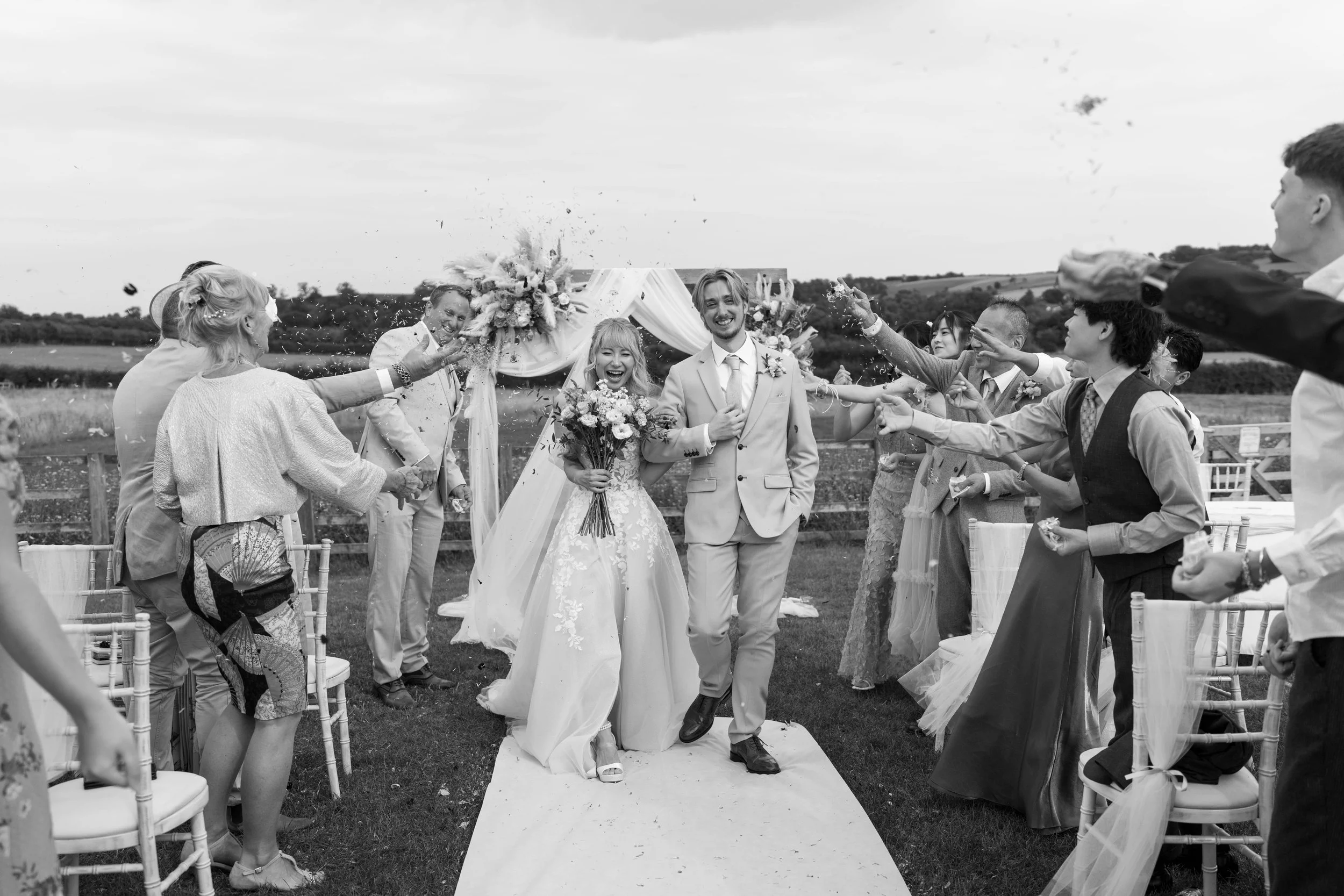 Black and white photo of a newly married couple walking down the aisle outside, surrounded by friends and family throwing confetti and celebrating their wedding.