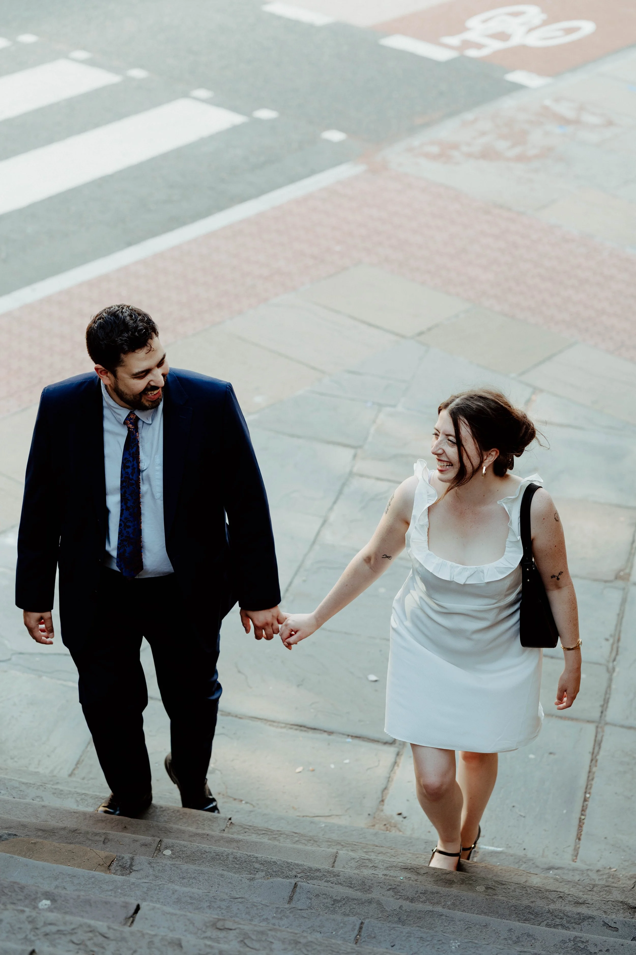 A couple holding hands and smiling as they walk up outdoor stairs, with a street and crosswalk visible in the background.