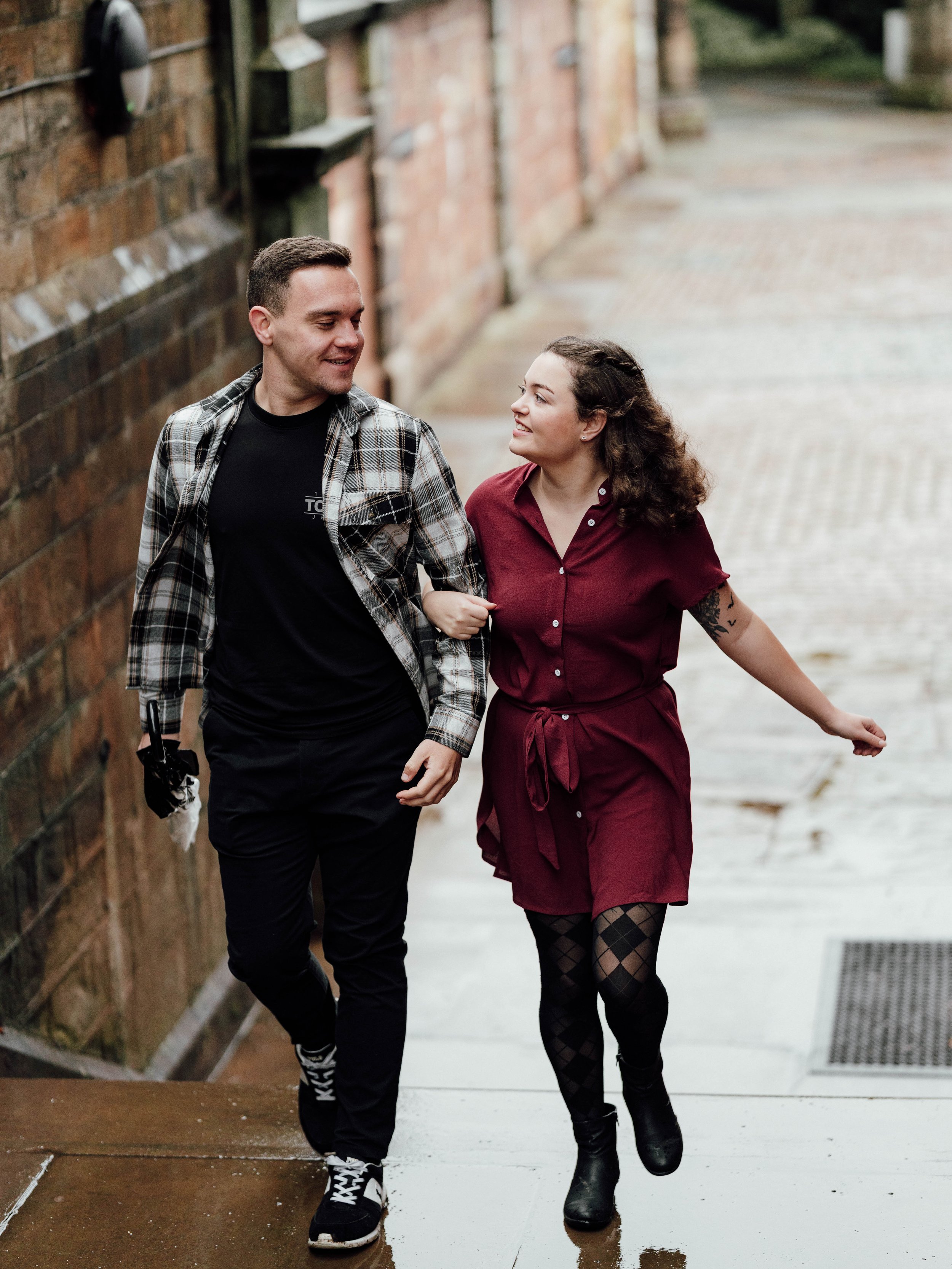 A young man and woman walk arm-in-arm on a wet sidewalk. The man is wearing a black t-shirt, black pants, sneakers, and a plaid shirt. The woman is dressed in a burgundy dress, black patterned tights, and boots. They are smiling and looking at each o