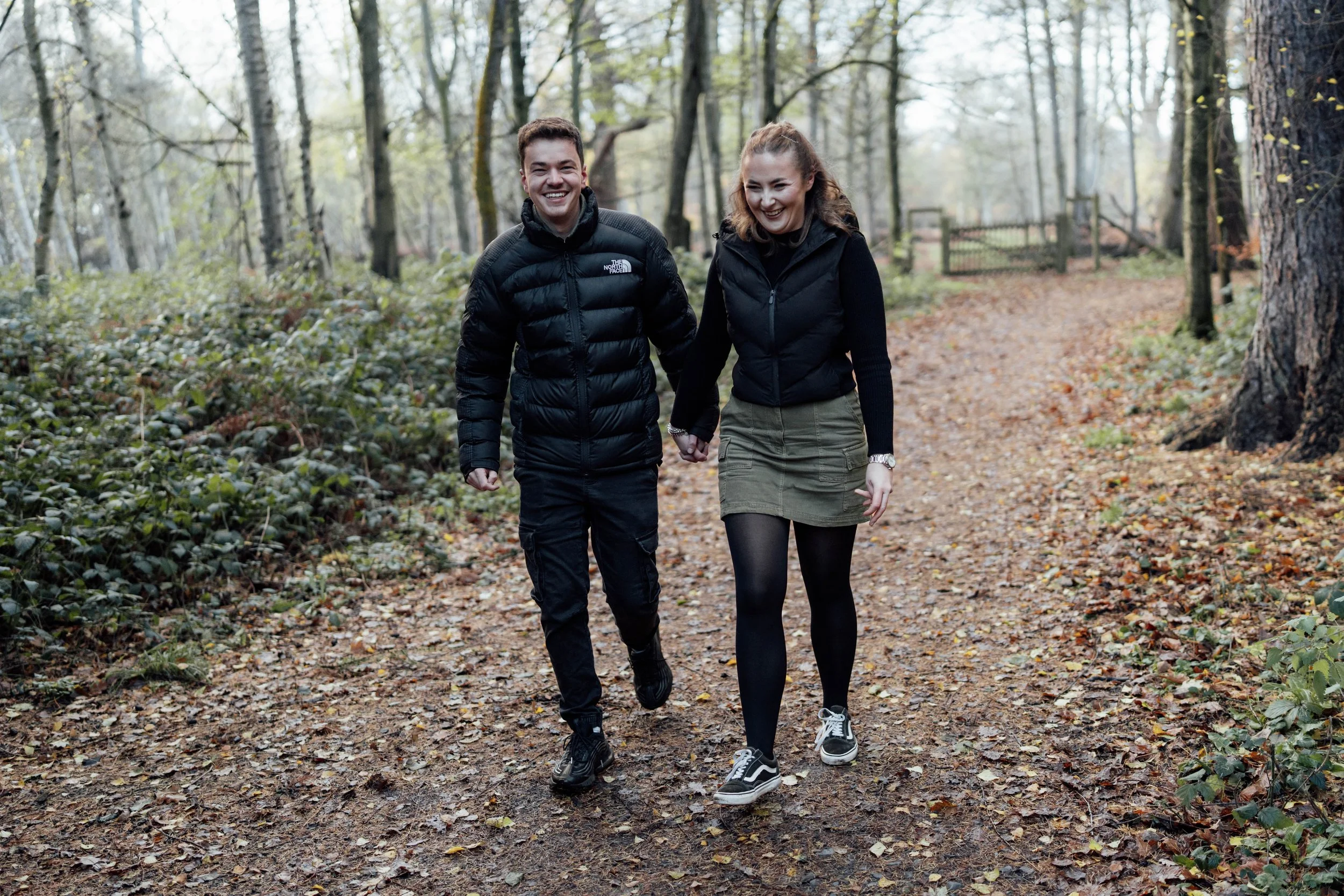 A young couple walking hand-in-hand on a forest trail during autumn, smiling and wearing casual outdoor clothing.