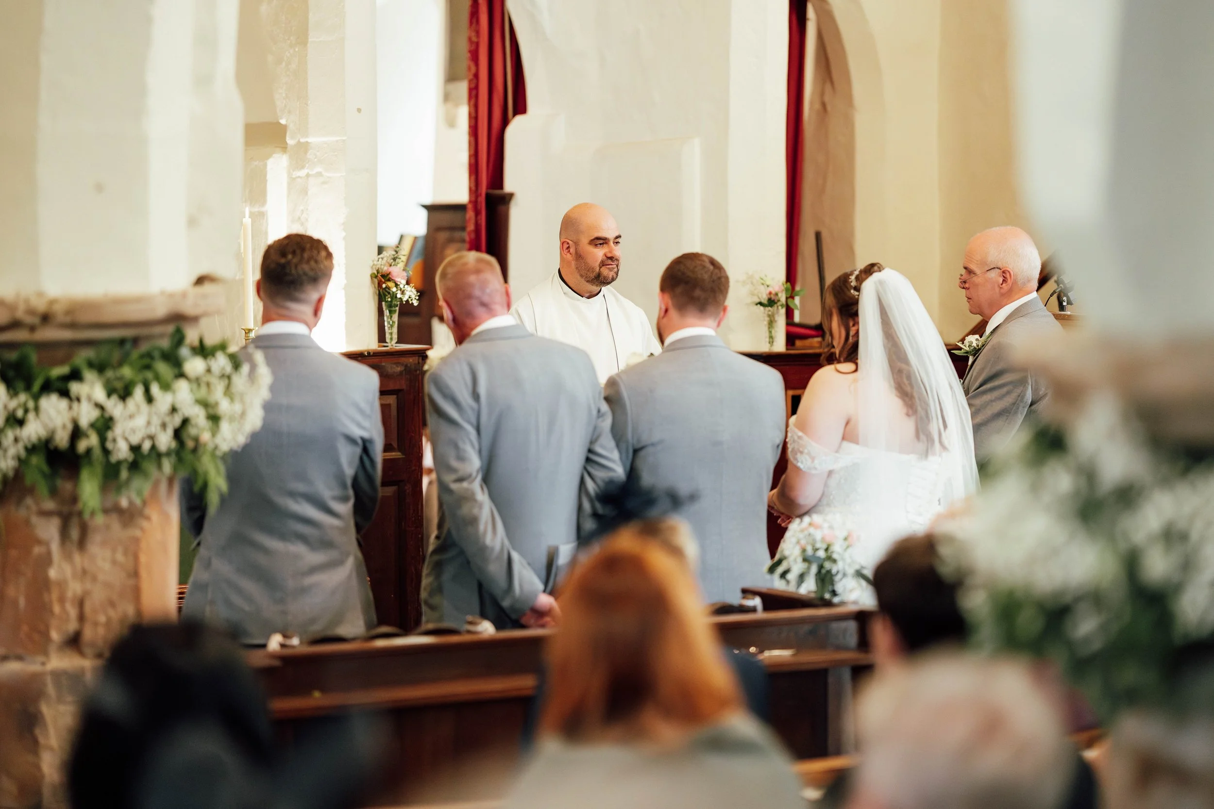 A wedding ceremony inside a church with six people standing at the altar, including a bride in a white gown with a veil and five men in suits, some with their backs to the camera and one facing forward.