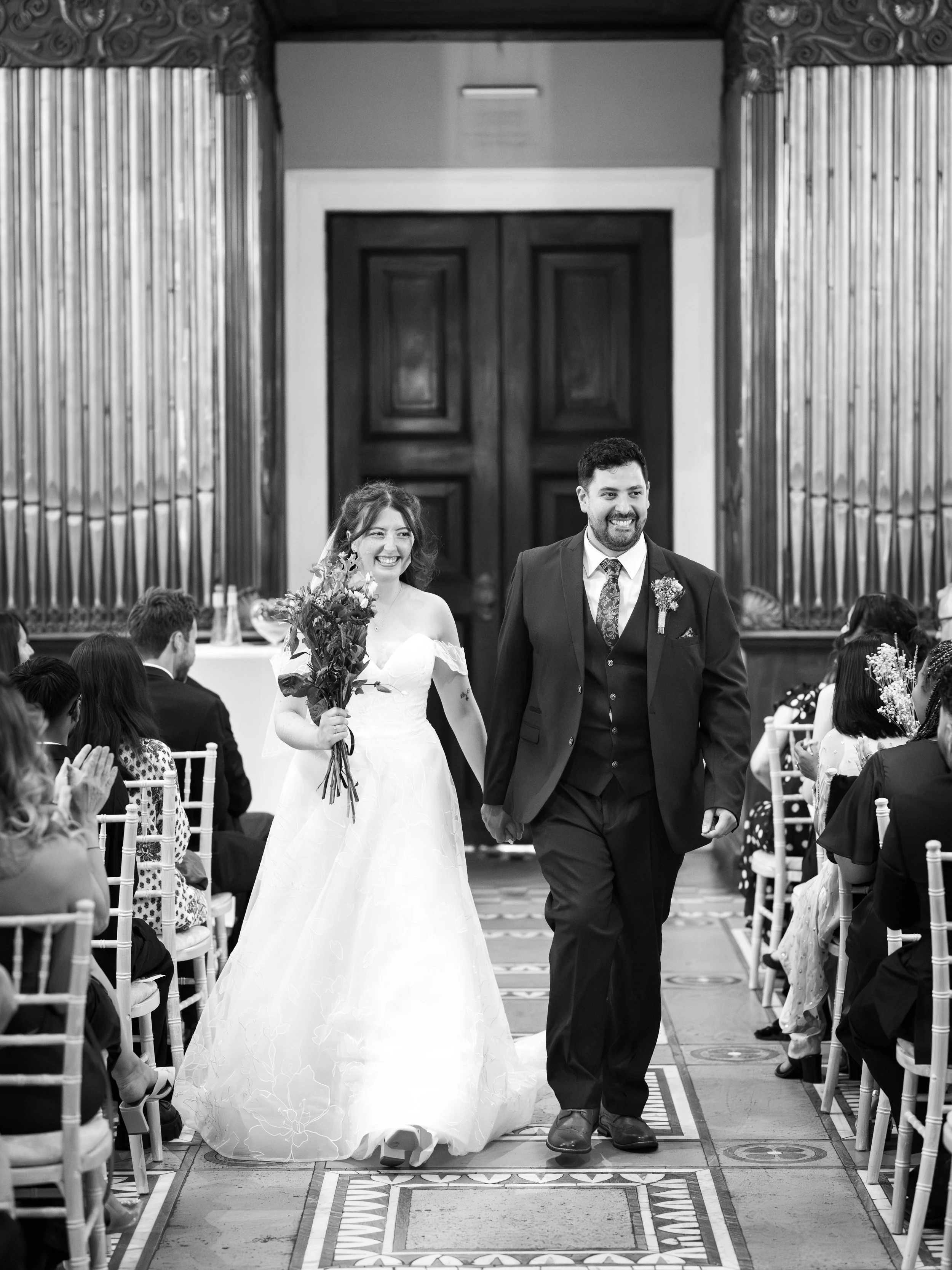 Black and white photo of a newlywed couple walking down the aisle after wedding ceremony, with wedding guests seated on either side.