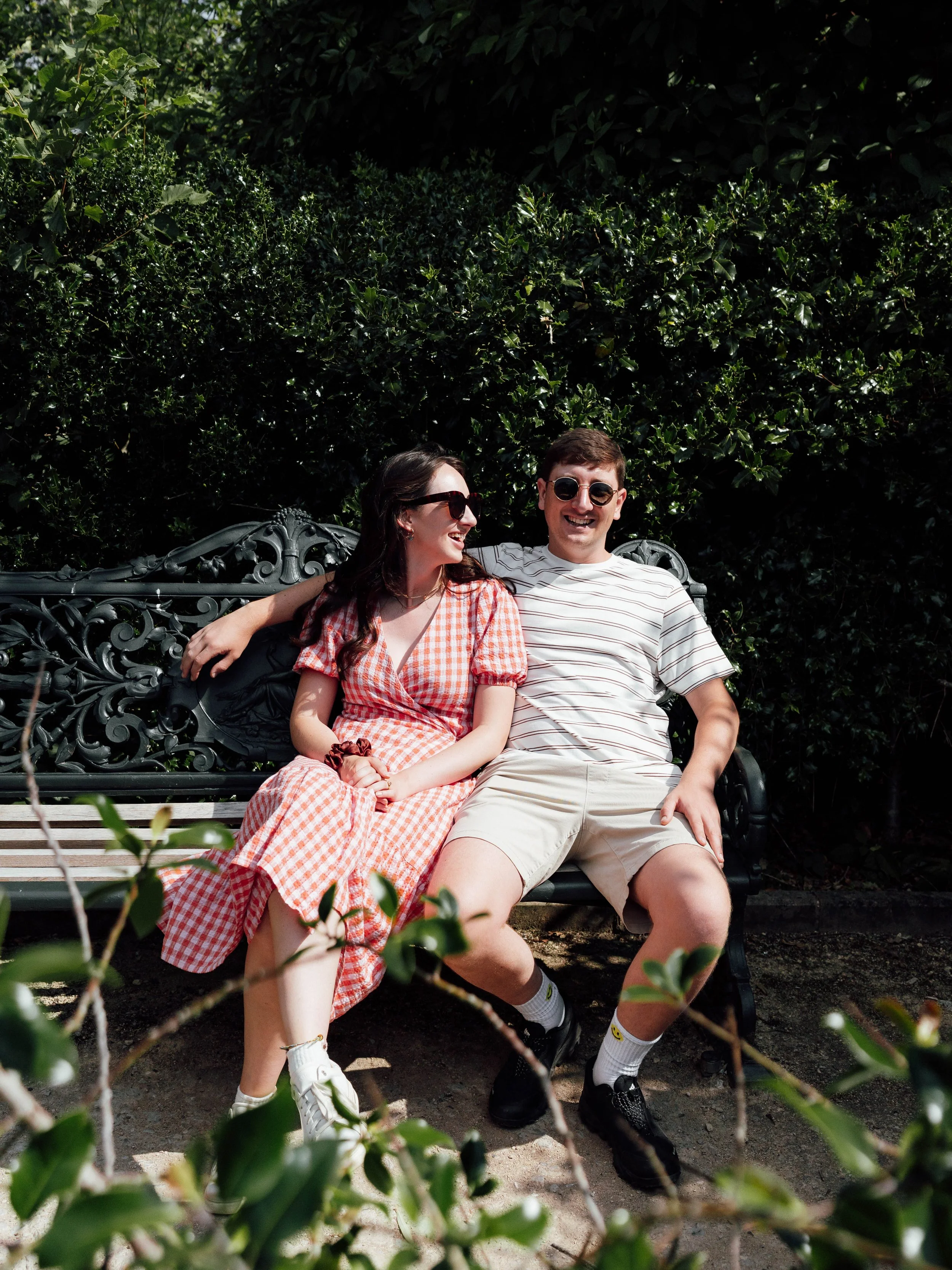A young woman and man sitting on a black ornate bench outdoors, smiling and enjoying each other's company, with green foliage in the background.