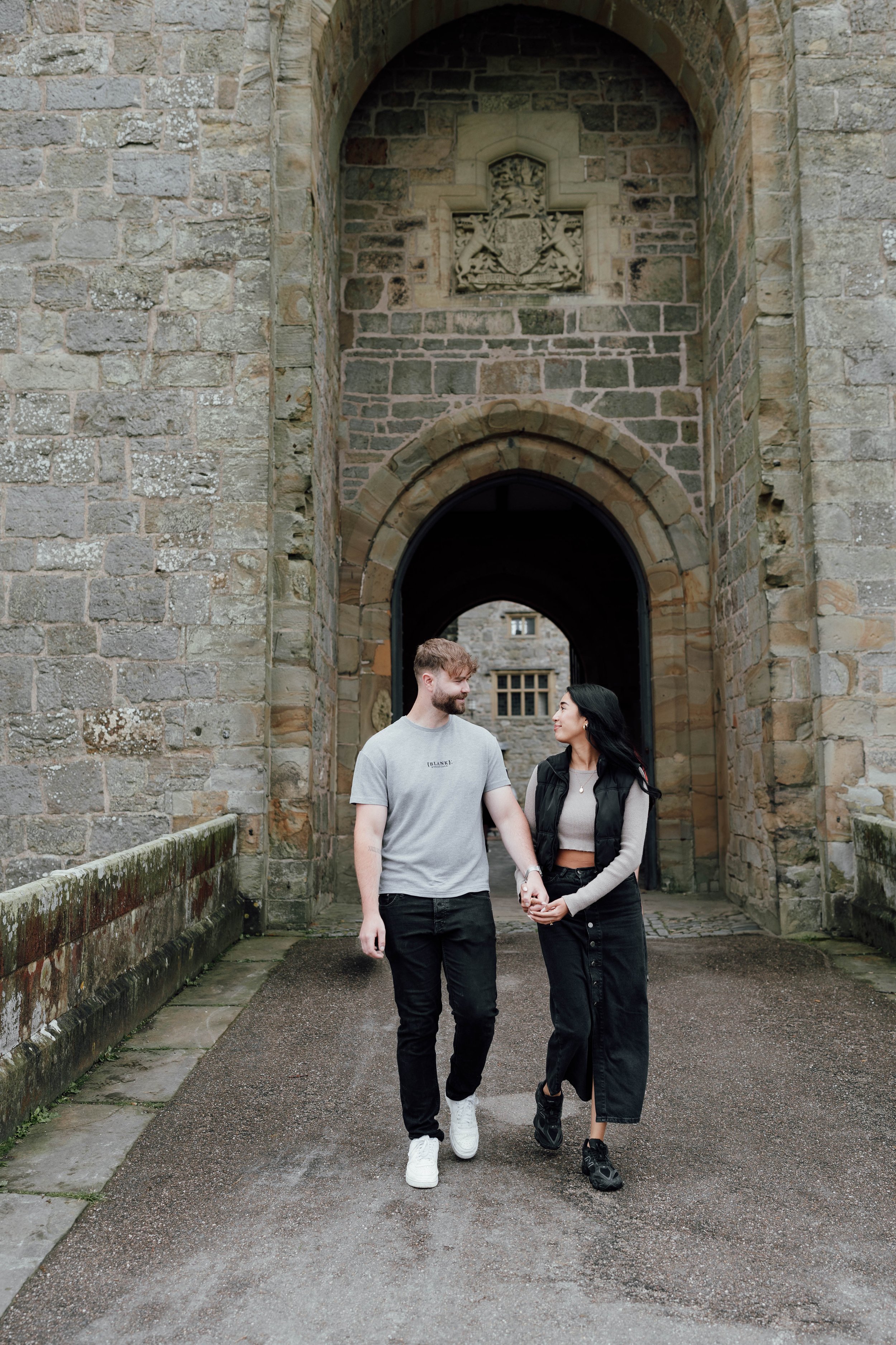 A young man and woman holding hands and walking through an arched stone gateway between old stone buildings, smiling at each other.