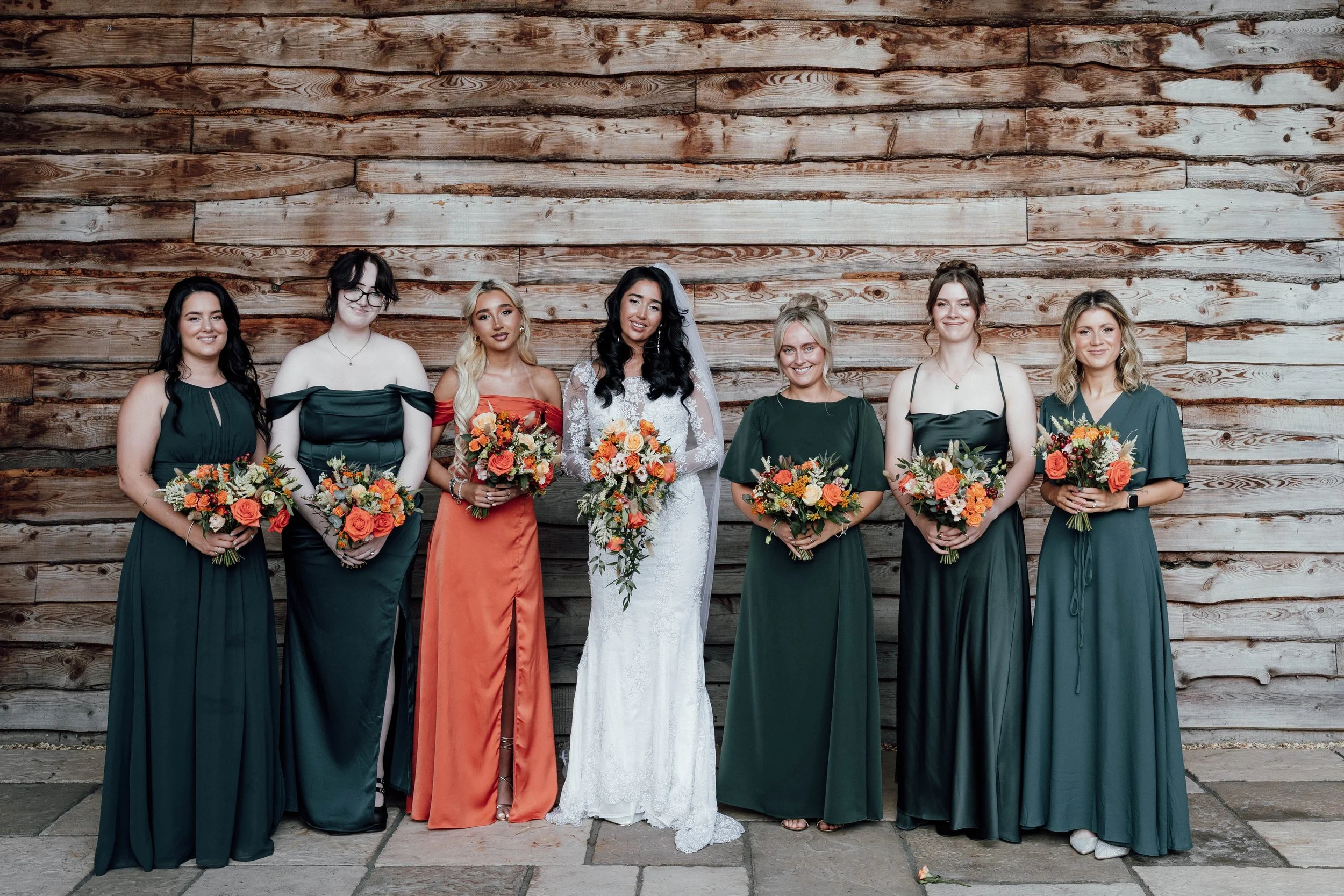 A bride and six bridesmaids standing in front of a wooden wall, each holding a bouquet of orange and peach flowers.