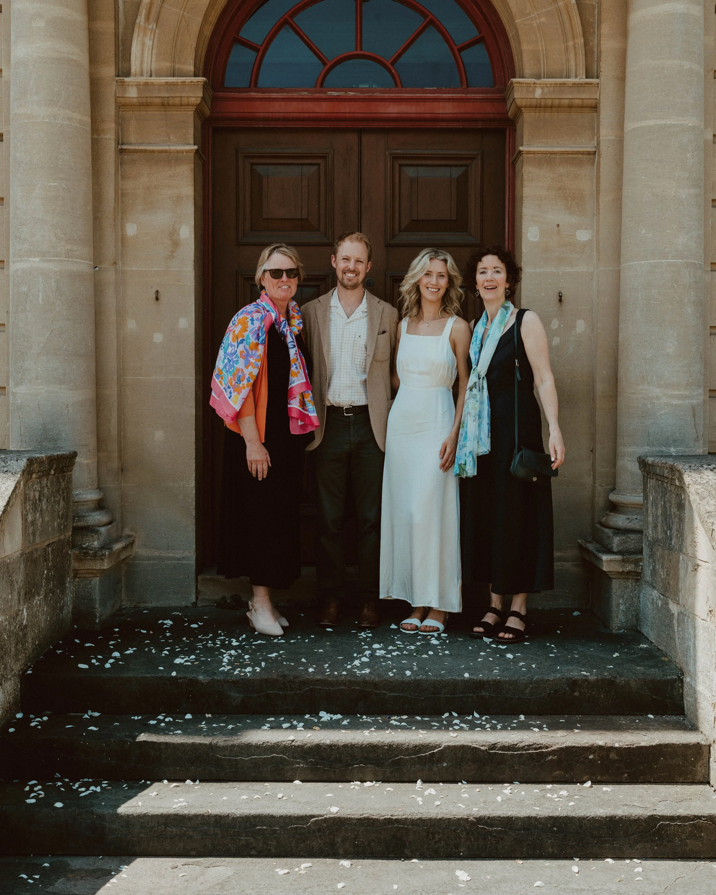 Four people standing on stone steps in front of a large wooden door, celebrating a wedding, with white flower petals scattered on the steps.