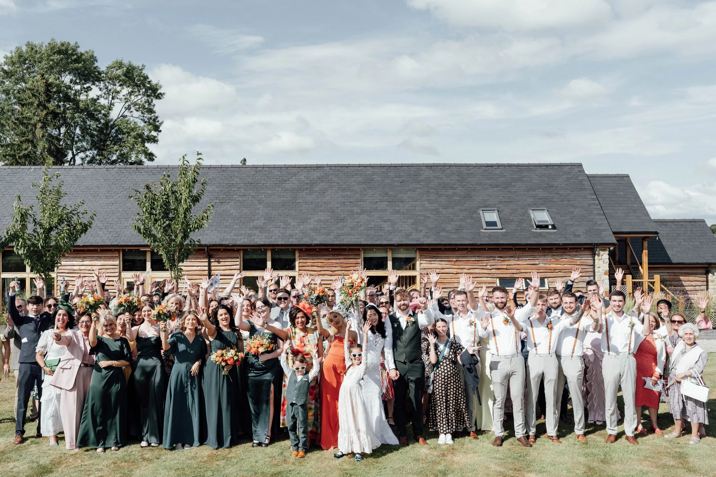 A large group of people at a wedding celebration outdoors, standing on grass in front of a rustic wooden building, with some holding bouquets, all raising their hands in the air, under a partly cloudy sky.