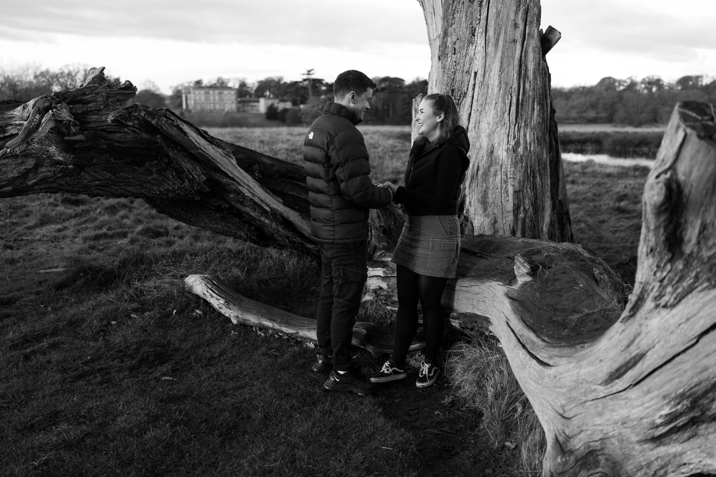 A happy couple stands holding hands and facing each other outdoors near a large fallen tree, with a wide field and a building in the background, in black and white.