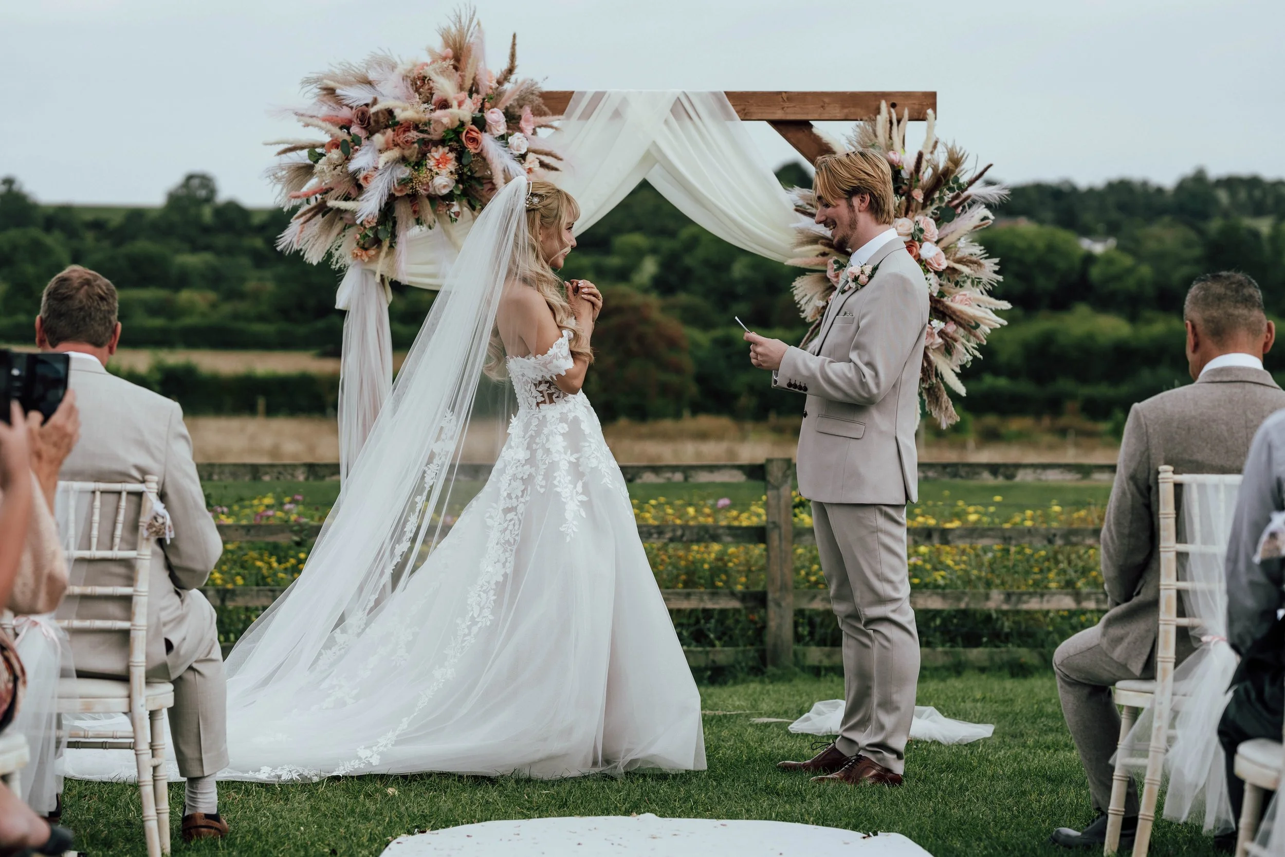 A couple getting married outdoors under a wooden arch decorated with flowers and draped fabric, with the bride in a white wedding gown and veil, and the groom in a light gray suit, exchanging vows.