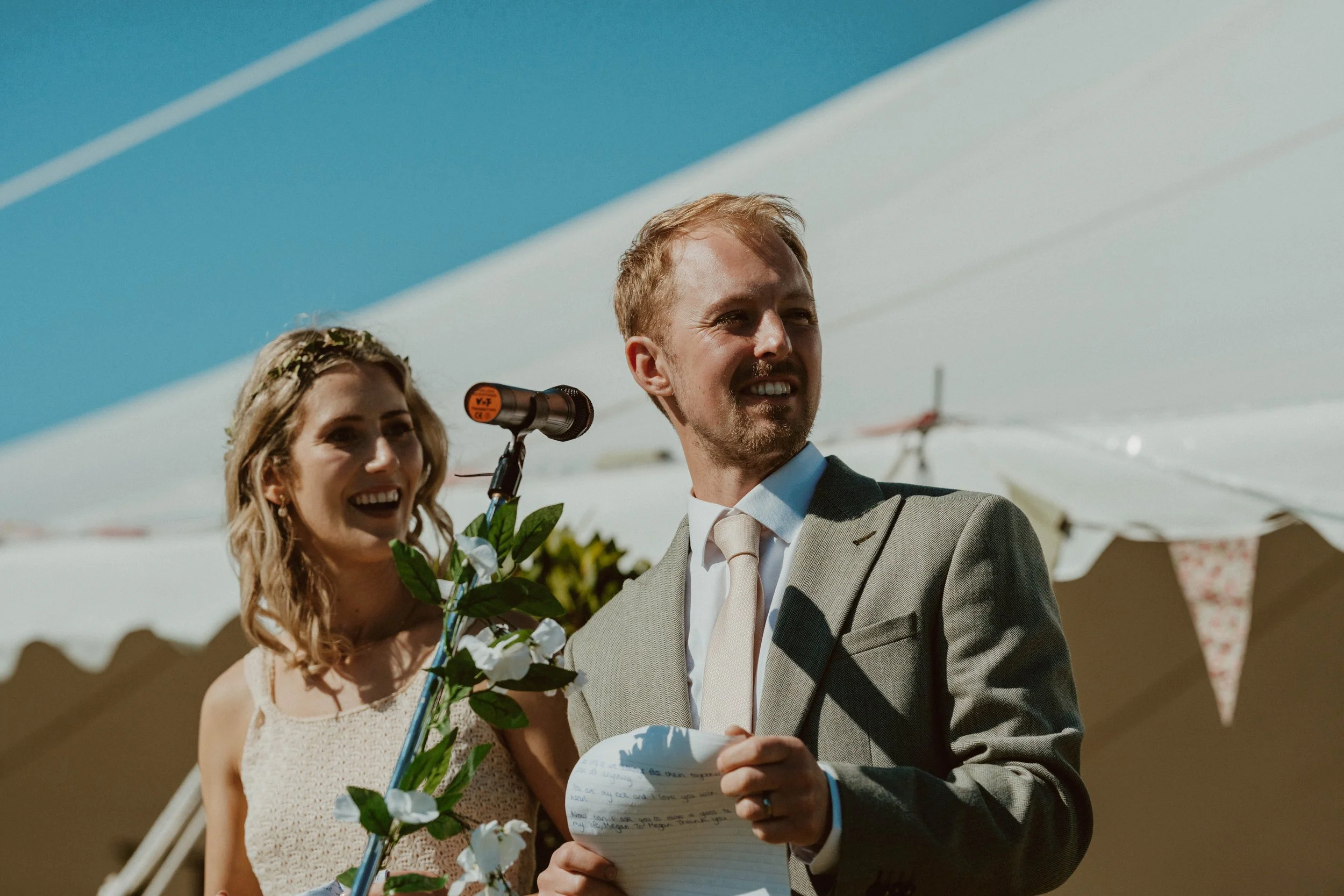 A bride and groom smiling at their outdoor wedding ceremony with a tent and bunting in the background.