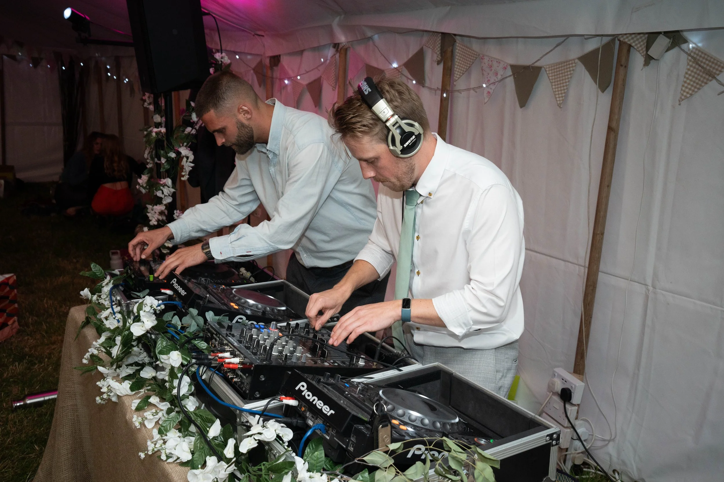 Two men DJing at a party, with sound equipment and floral decorations, inside a tent with string lights and bunting.