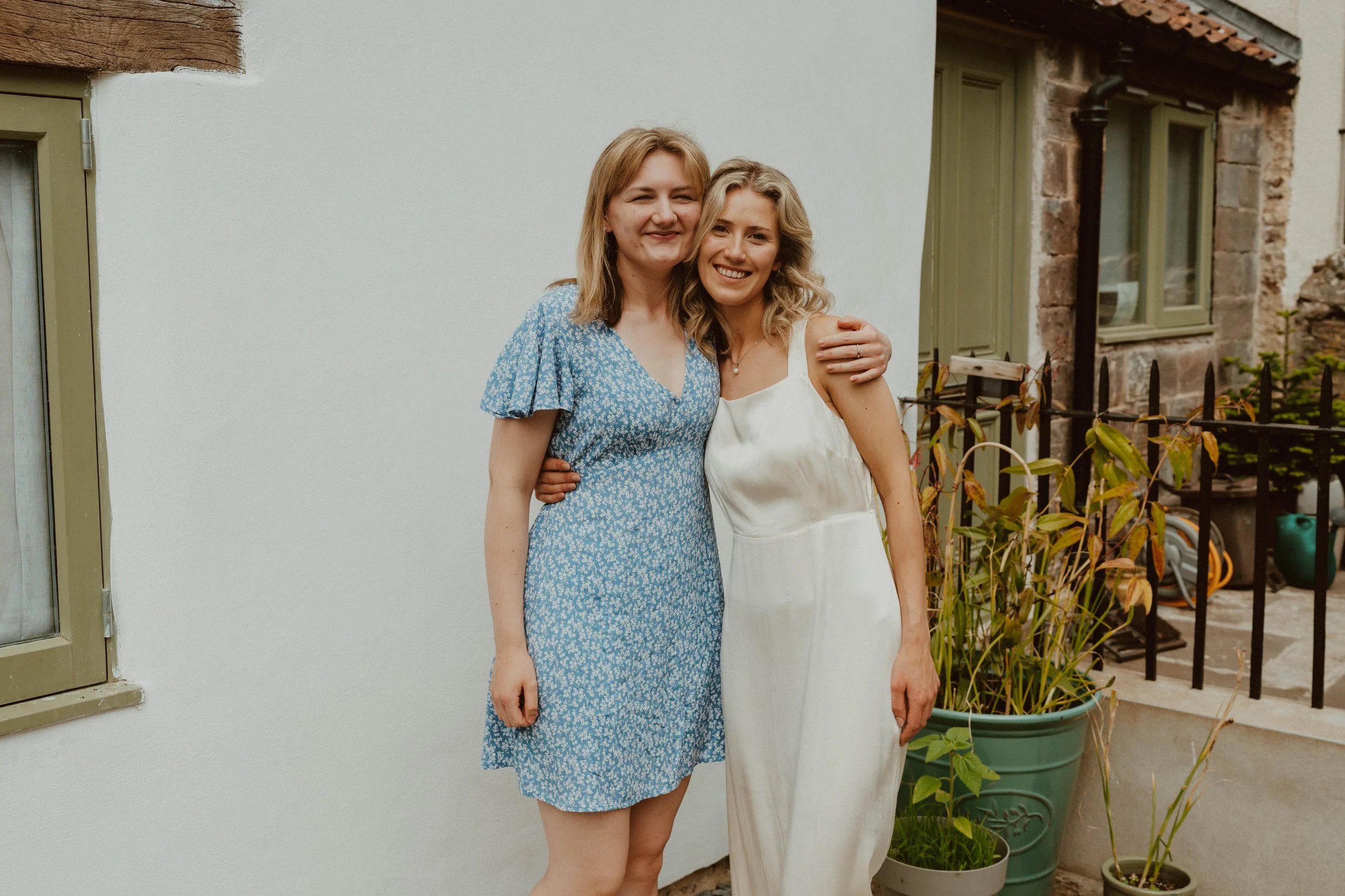 Two women standing close together outdoors, smiling at the camera. One is wearing a blue floral dress with short sleeves, and the other is in a ivory satin slip dress. There are potted plants and a black metal fence in the background.