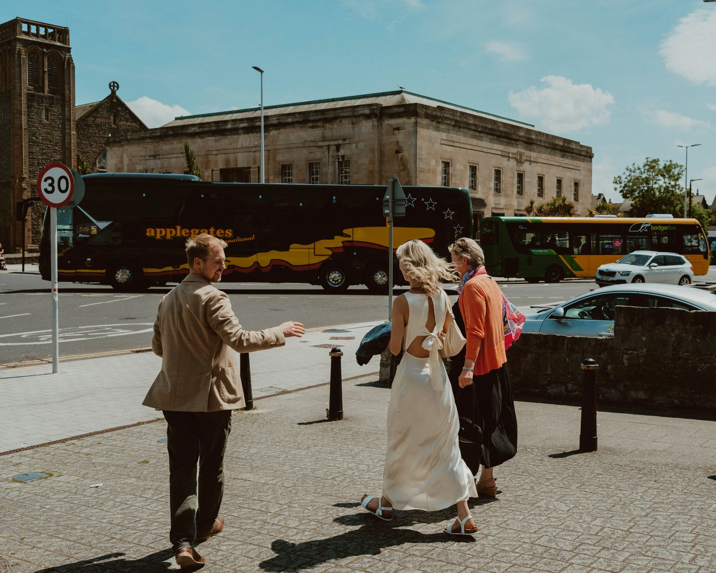 Three people standing on a sidewalk near a street, with a bus passing by in the background on a sunny day.