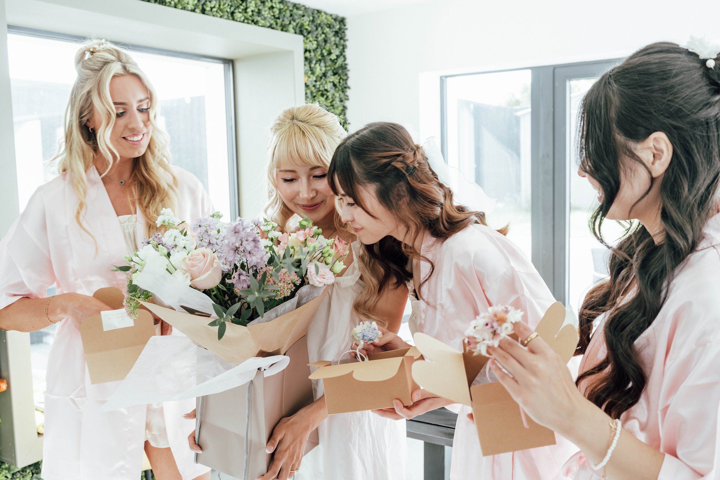 Group of women in pink robes exchanging flowers and gifts indoors.