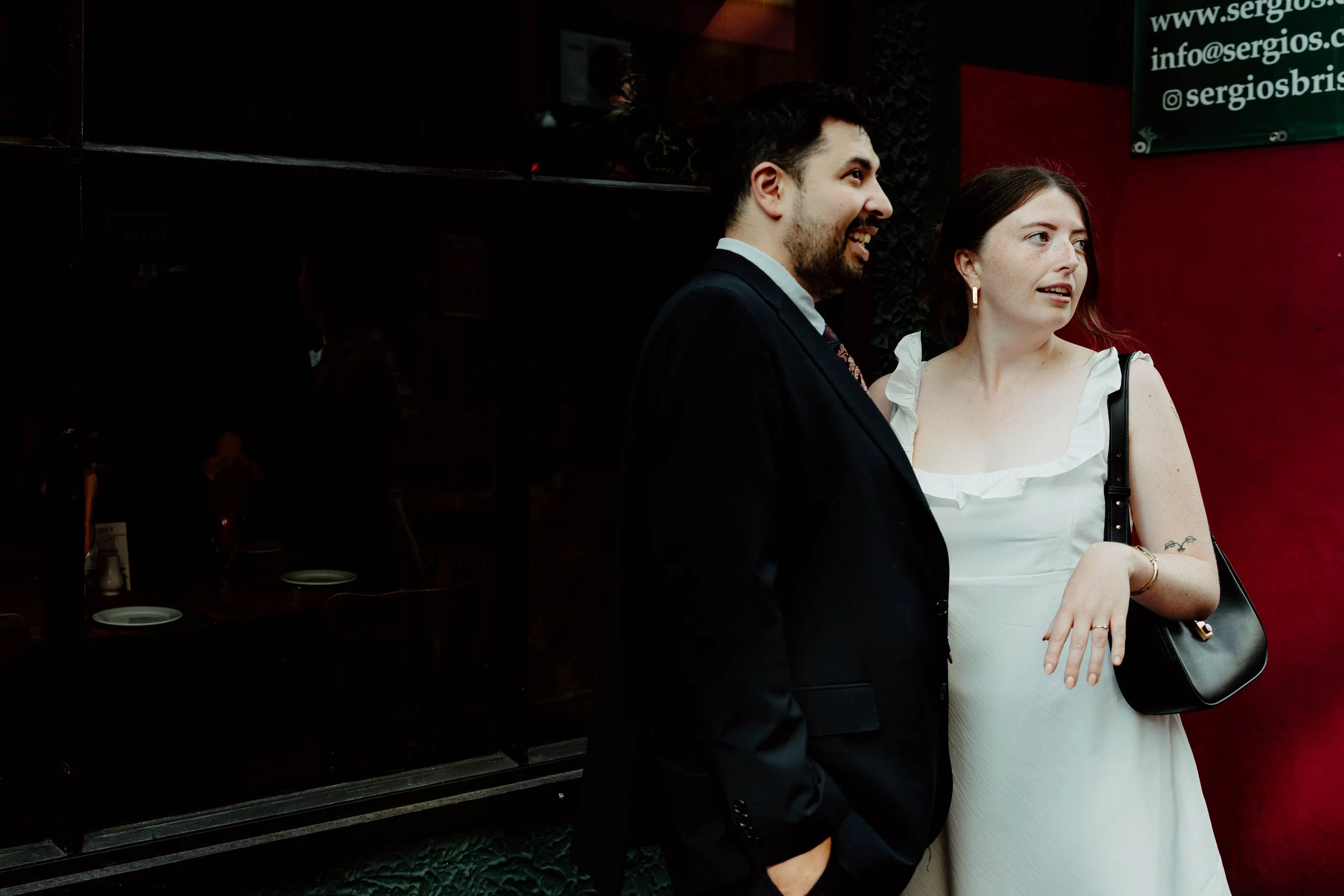 A man and a woman stand together outside a building, engaged in conversation. The man, wearing a dark suit, is smiling and facing left. The woman, dressed in a white dress with ruffled sleeves, has a black purse over her shoulder and is looking to he