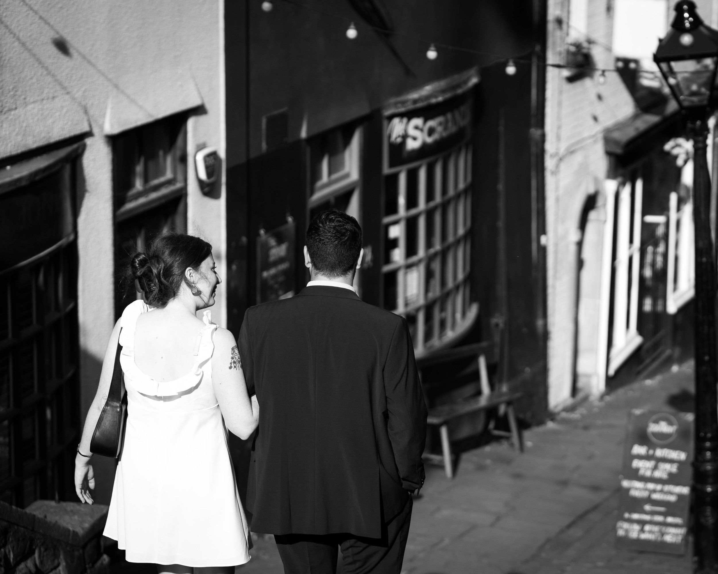 A woman and a man walk together on a city sidewalk at night. The woman has dark hair tied back, is wearing a white dress with ruffled straps, and has a tattoo on her upper arm. The man is dressed in a dark suit with short hair. They are photographed 