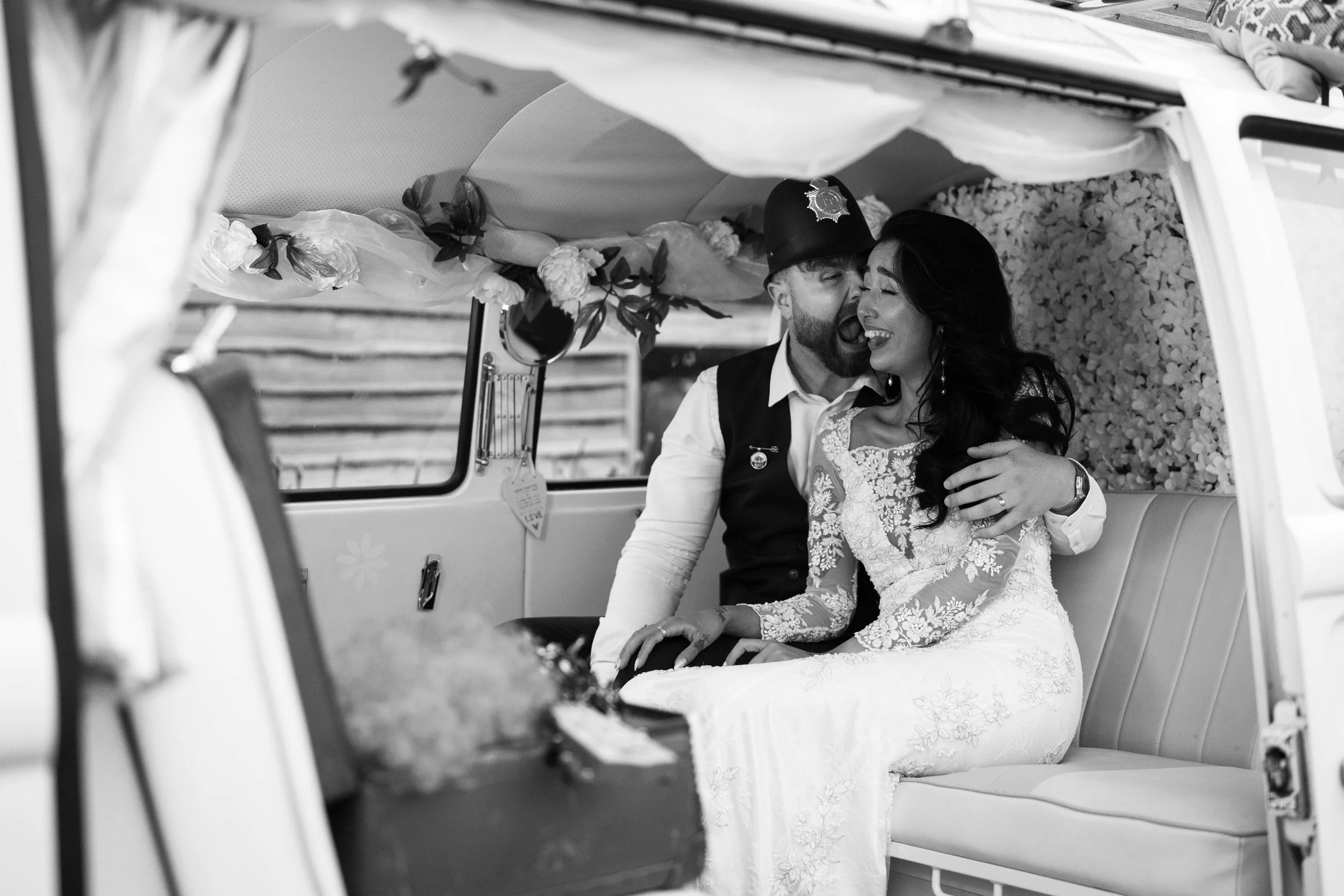 Black and white photo of a bride and groom in a decorated van, smiling and hugging. The bride wears a lace wedding dress, and the groom is dressed as a police officer. The van's interior is decorated with flowers and fabric.