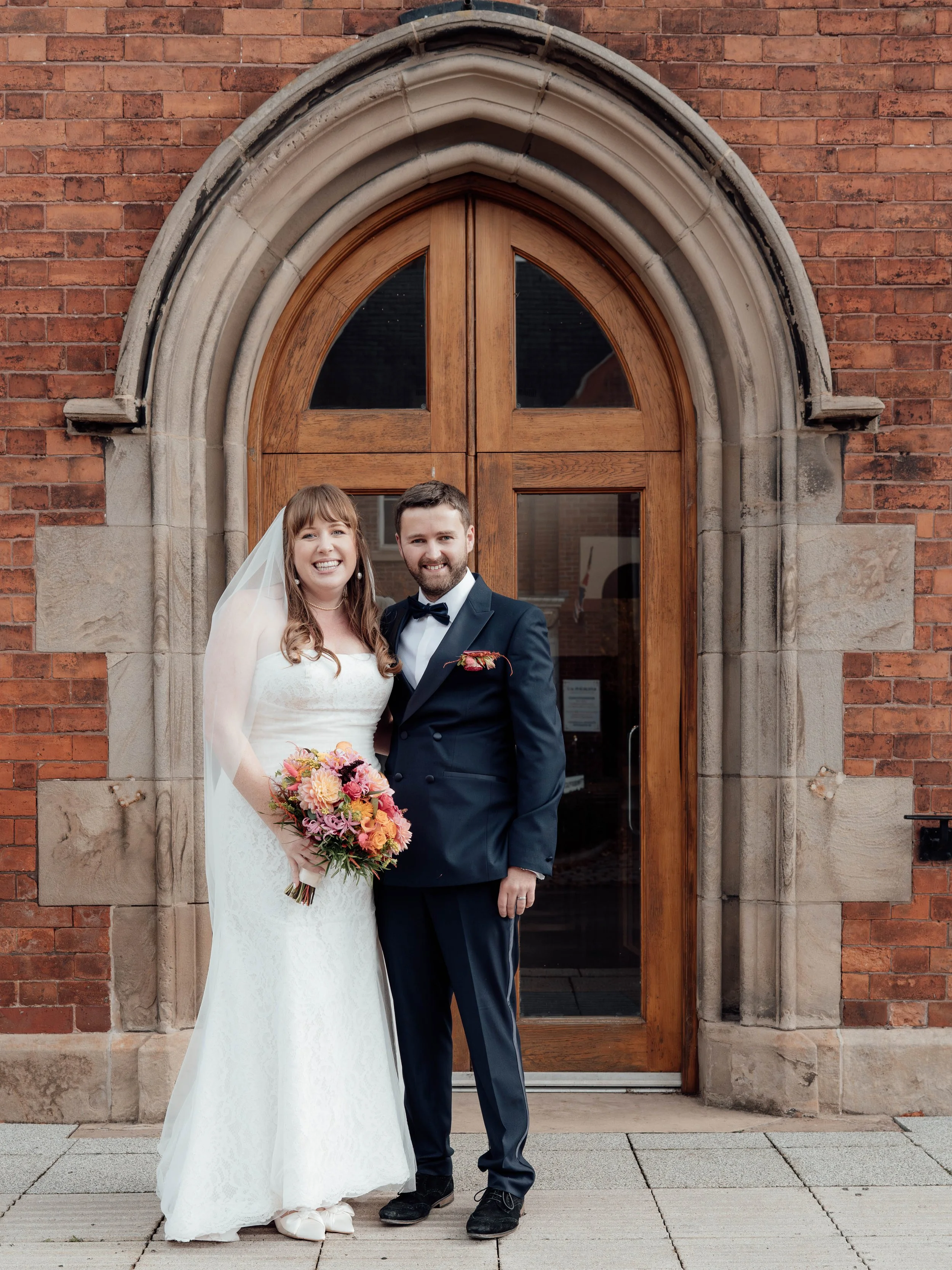 A bride and groom standing in front of a wooden arched double door with brick walls surrounding it. The bride is dressed in a white wedding gown holding a colorful bouquet, and the groom is wearing a dark suit with a bow tie.