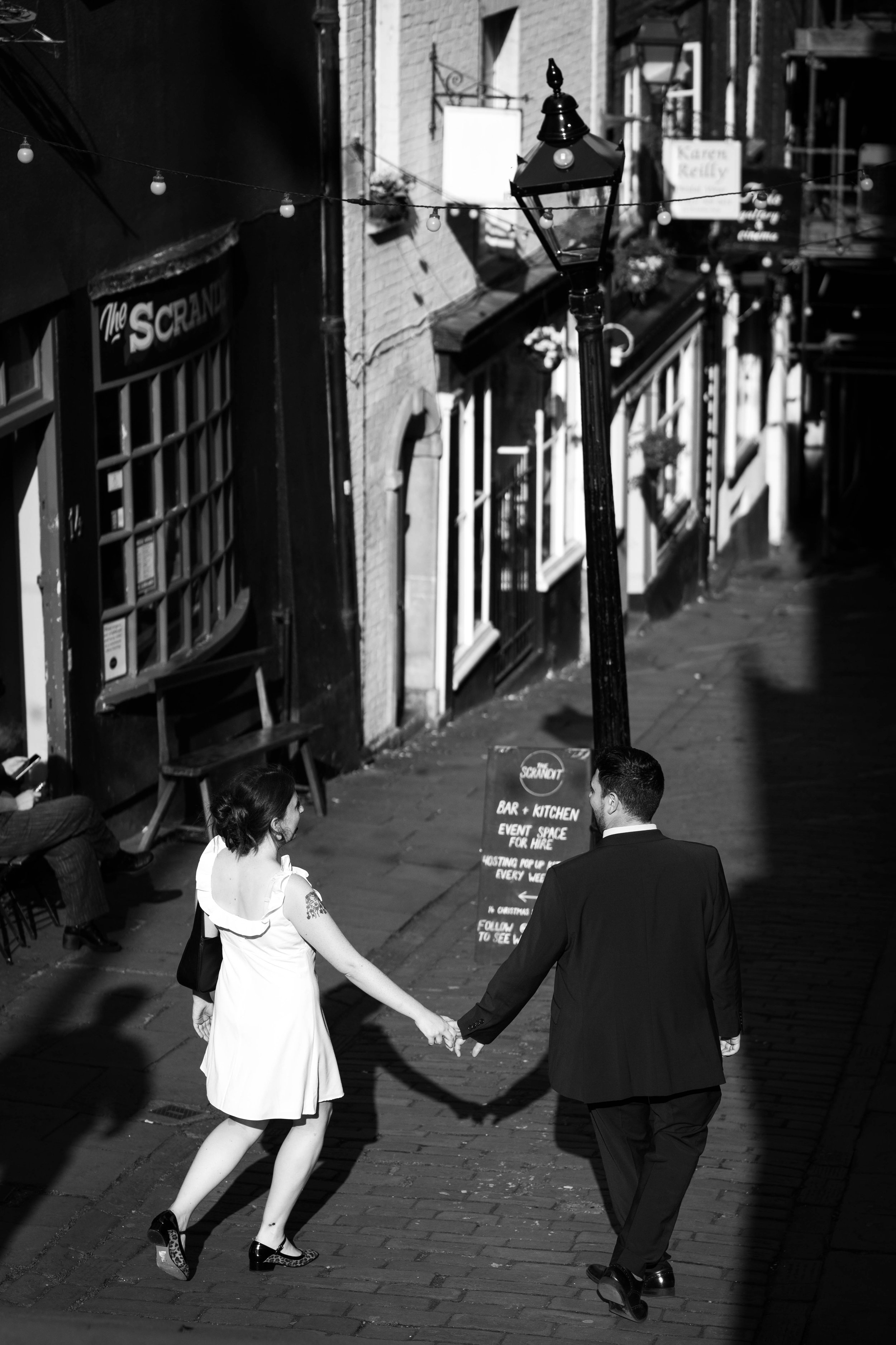 A couple holding hands and walking down a city street in black and white, with a woman wearing a white dress and a man in a dark suit. There are storefronts, a lamppost, and a sign in the background.