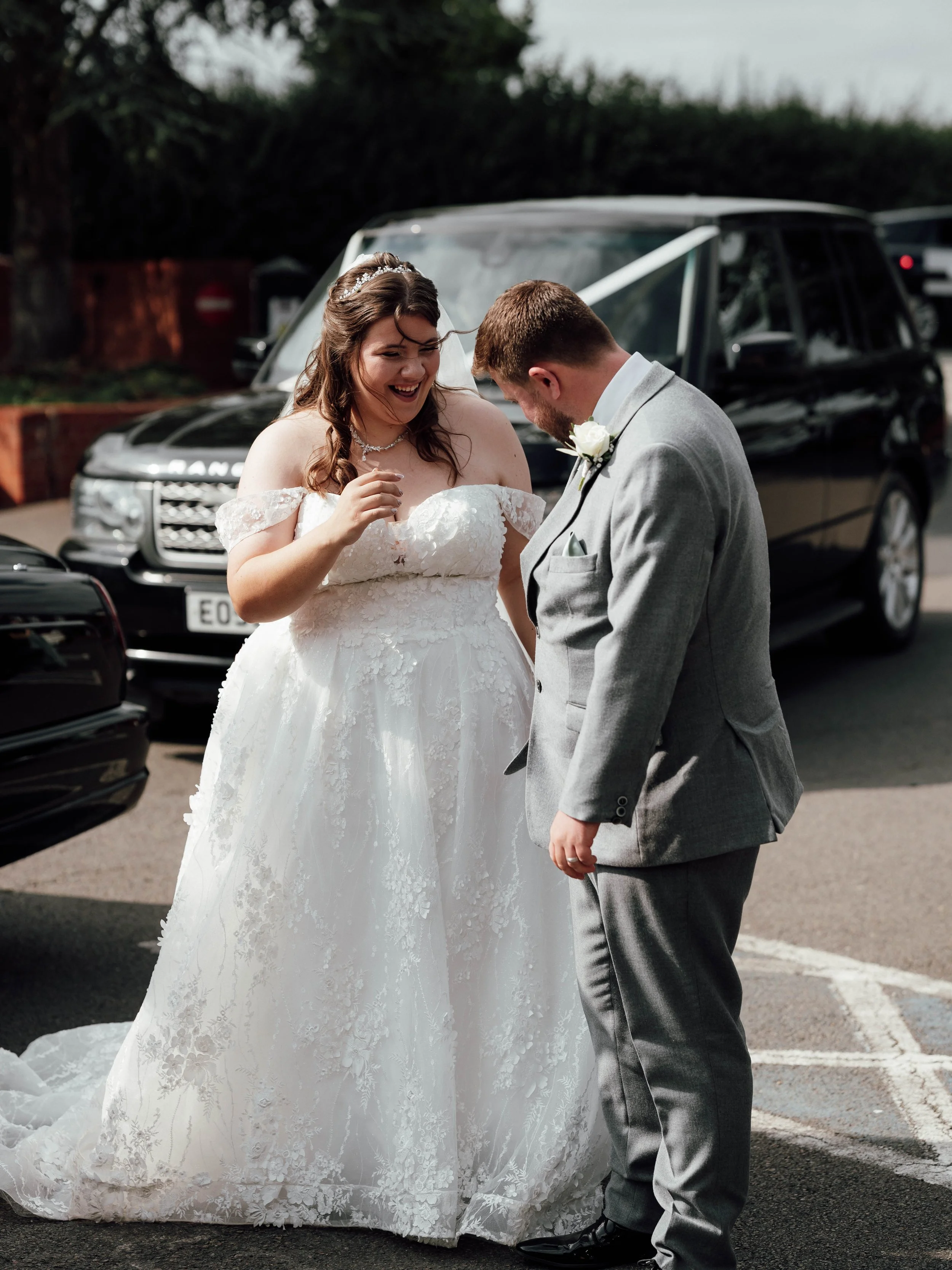 A bride and groom, dressed in wedding attire, share a joyful moment outside on a parking lot with cars in the background.