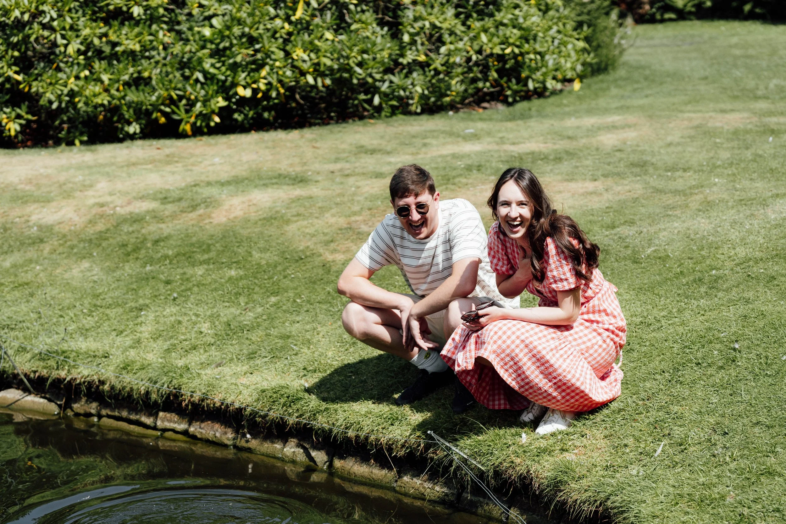 A young man and woman sitting on a grassy area near a pond, laughing and smiling together.