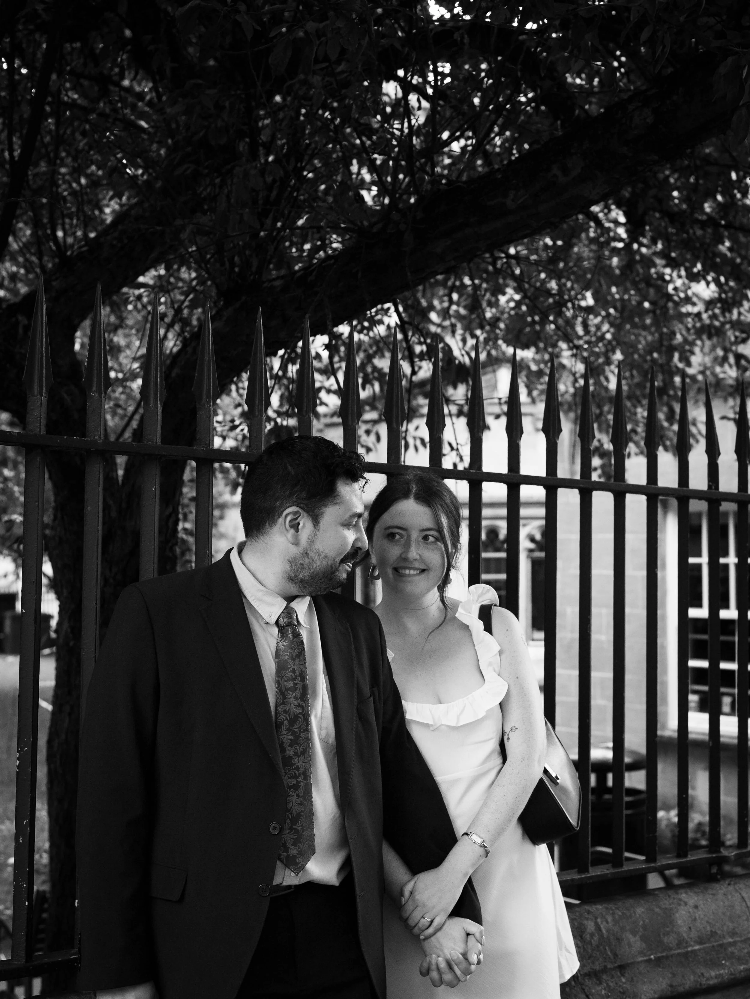A black and white photo of a couple standing close together, holding hands, outdoors in front of a fence and trees. The man is wearing a suit and the woman is wearing a dress with ruffles, both looking at each other.