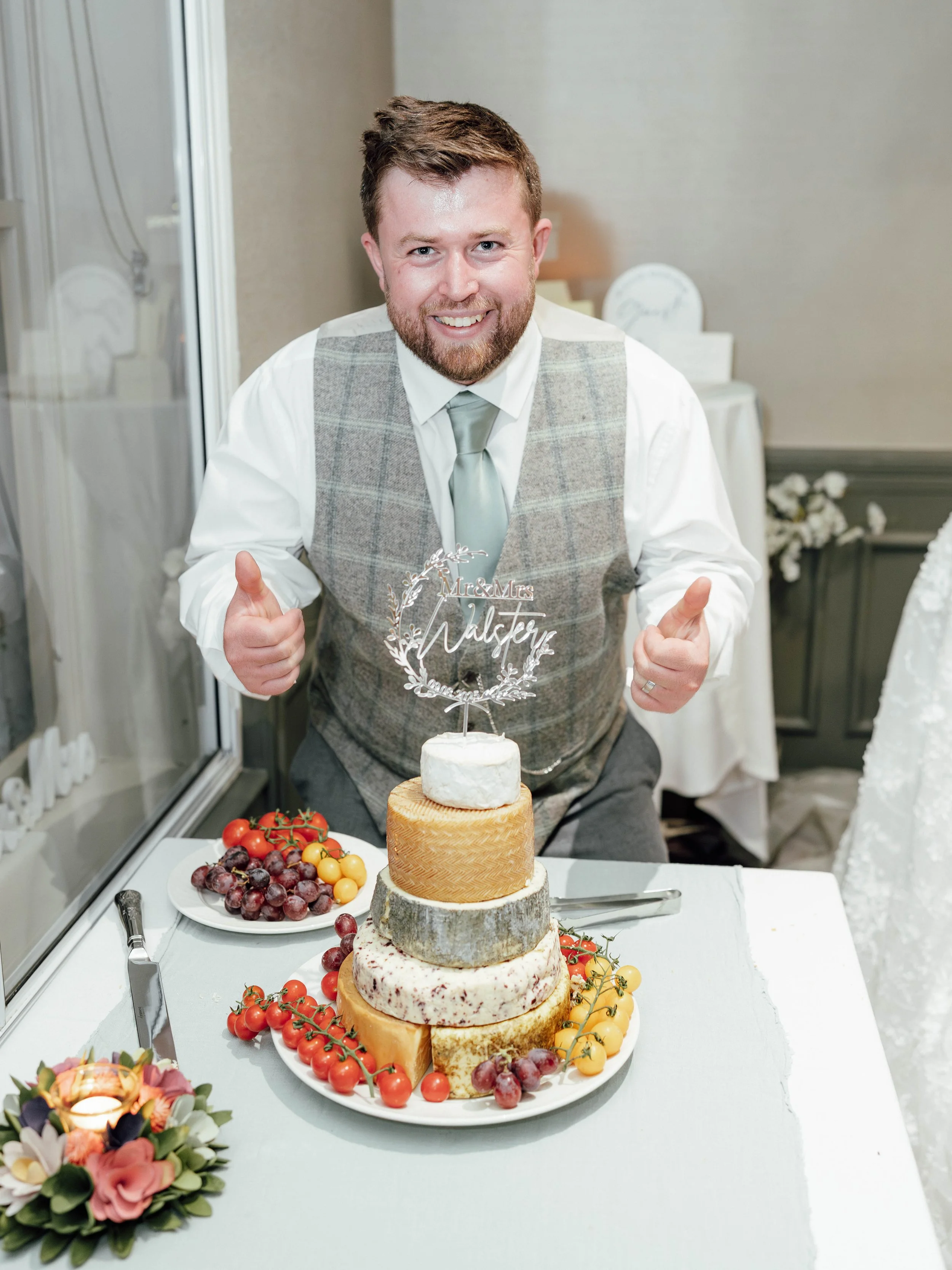 Smiling man in wedding attire giving thumbs up behind a wedding cake decorated with various cheeses and fruits, and a topper that reads 'Mr & Mrs Mulberry' at a wedding reception.
