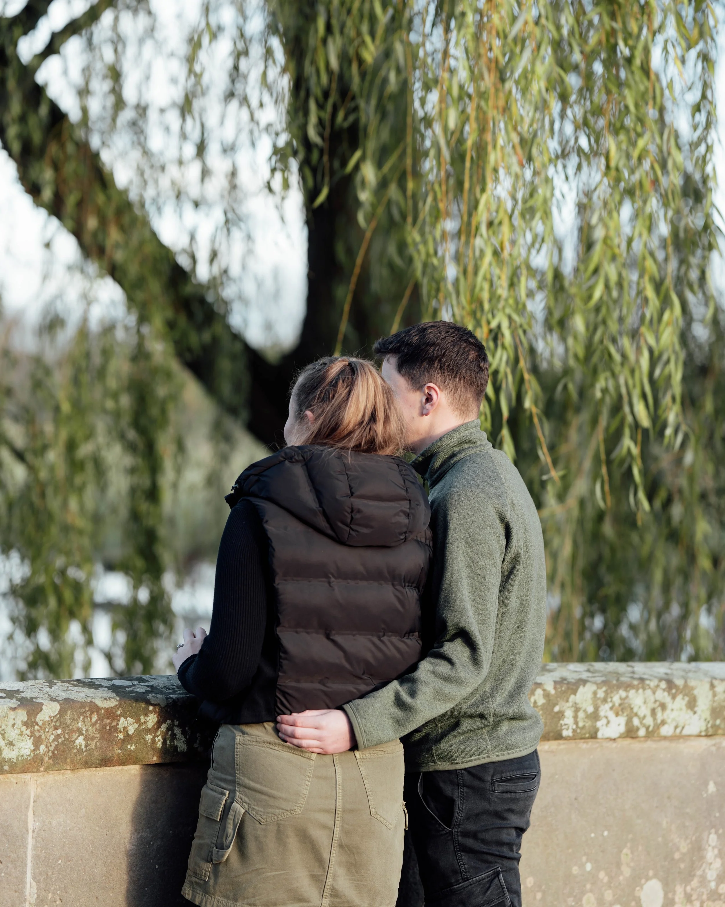 A young couple standing close and sharing a kiss by a stone wall with a large tree and hanging branches in the background.