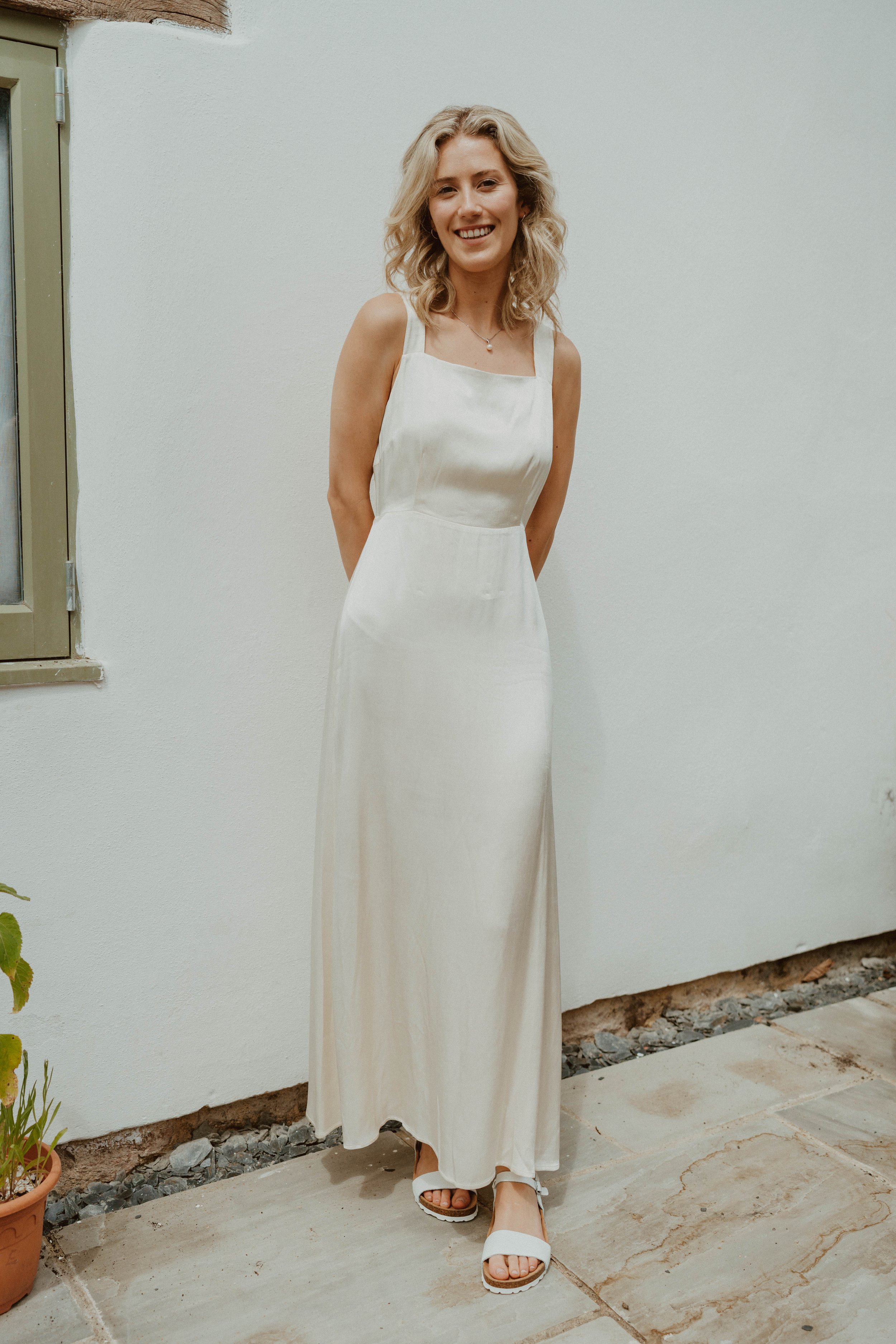 A young woman with blonde wavy hair wearing a white satin dress with straps, standing outdoors against a white wall, smiling at the camera.