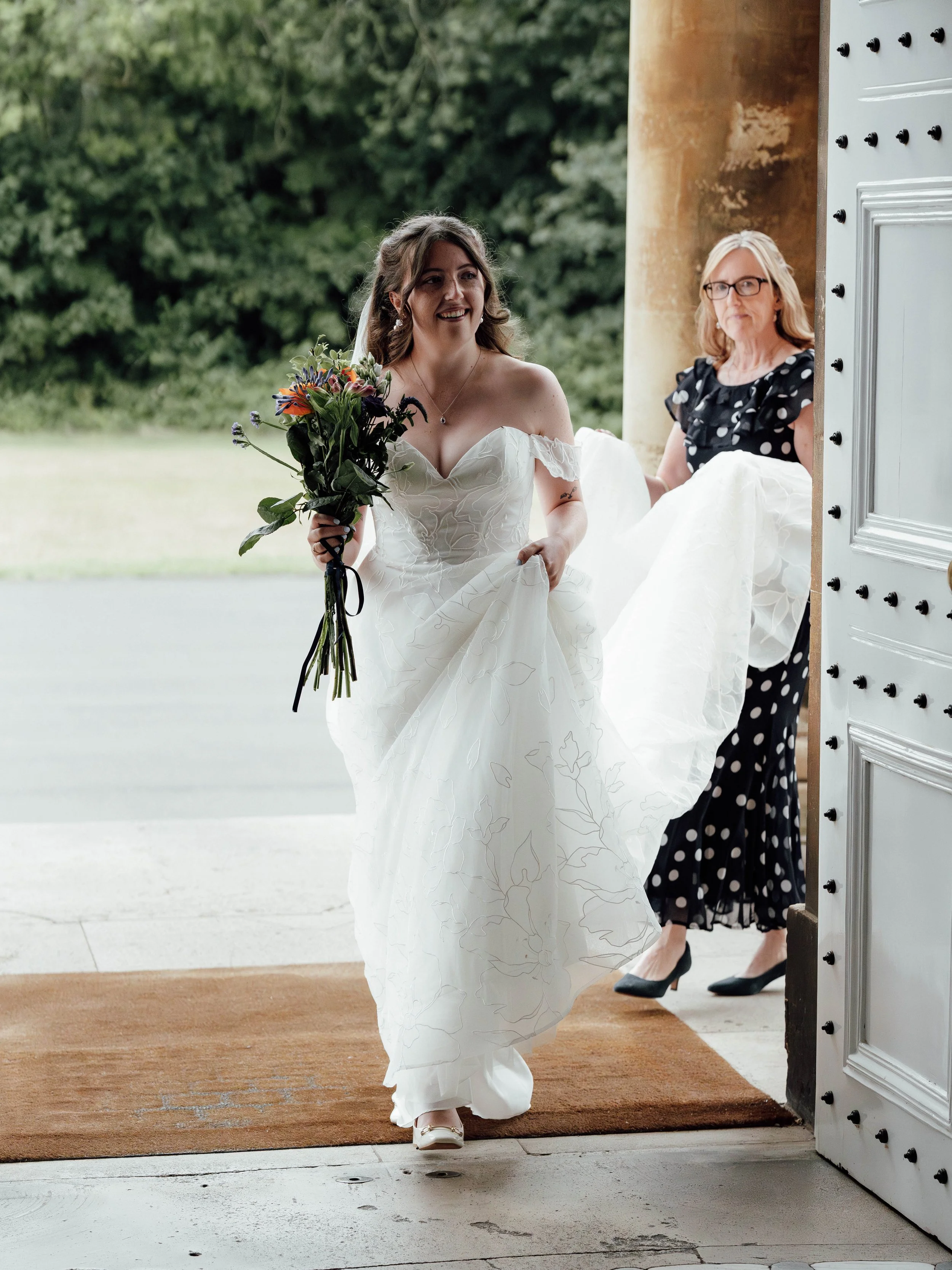 A bride in a white wedding dress holding a bouquet of flowers, smiling as she enters a building, with a woman in a black polka dot dress behind her.