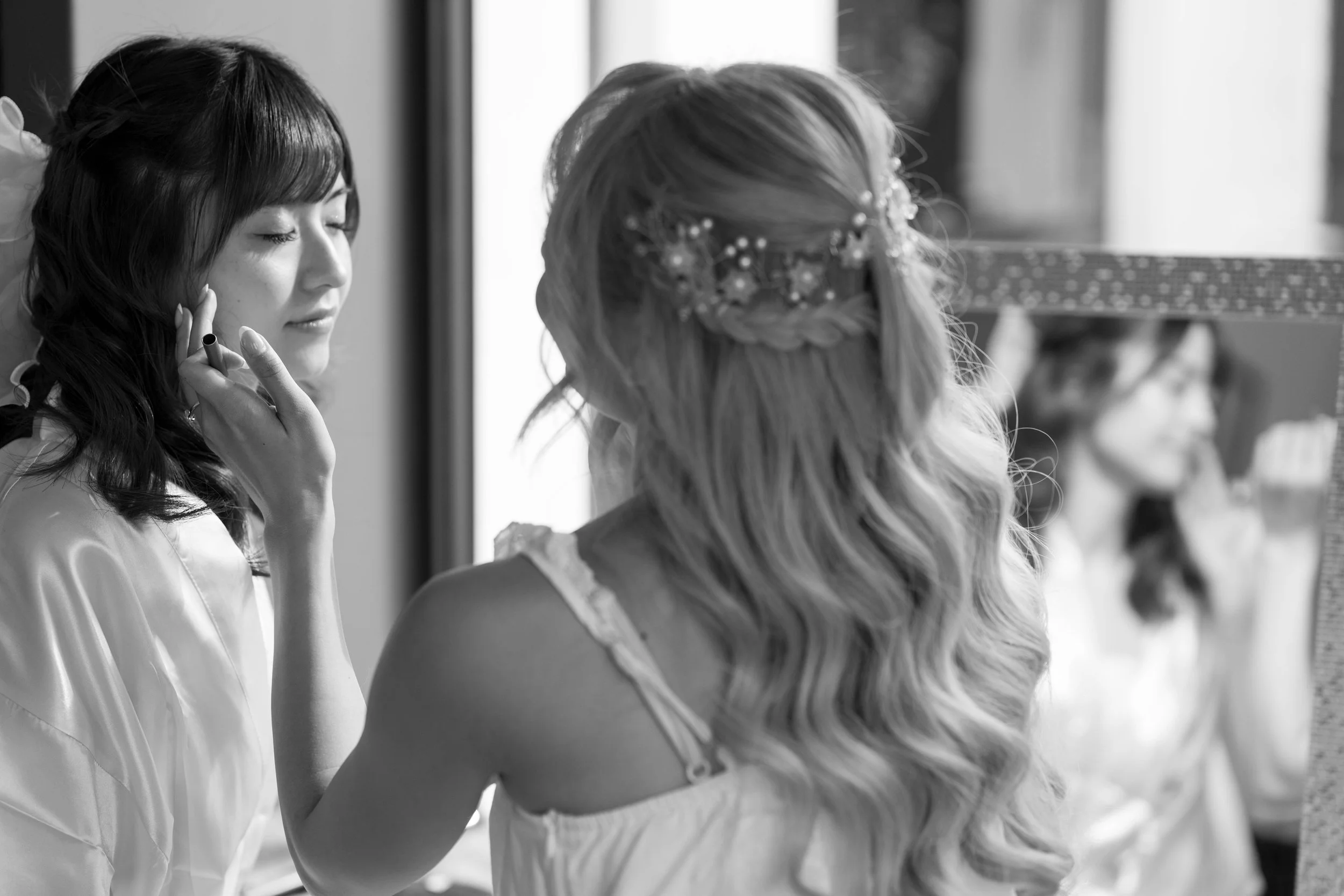 Two women, including a bride with long wavy hair and a decorative hairpiece, are looking in a mirror while one adjusts her makeup or hair. The scene is in black and white and appears to be preparations for a wedding.