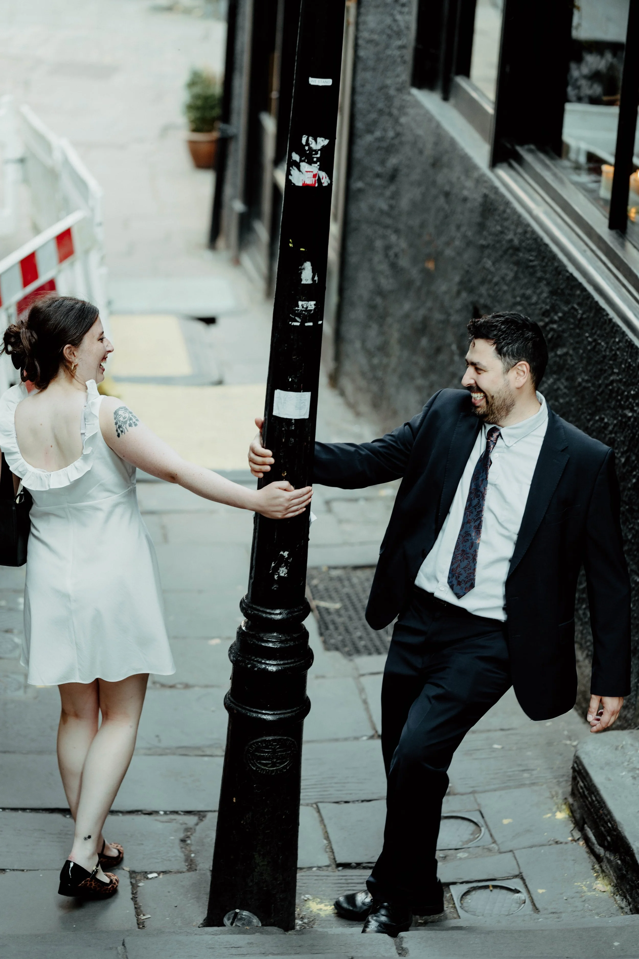 A man and woman are playing cheerfully on a city sidewalk, with the man pushing a black street pole while the woman reaches out to him, both smiling and laughing.