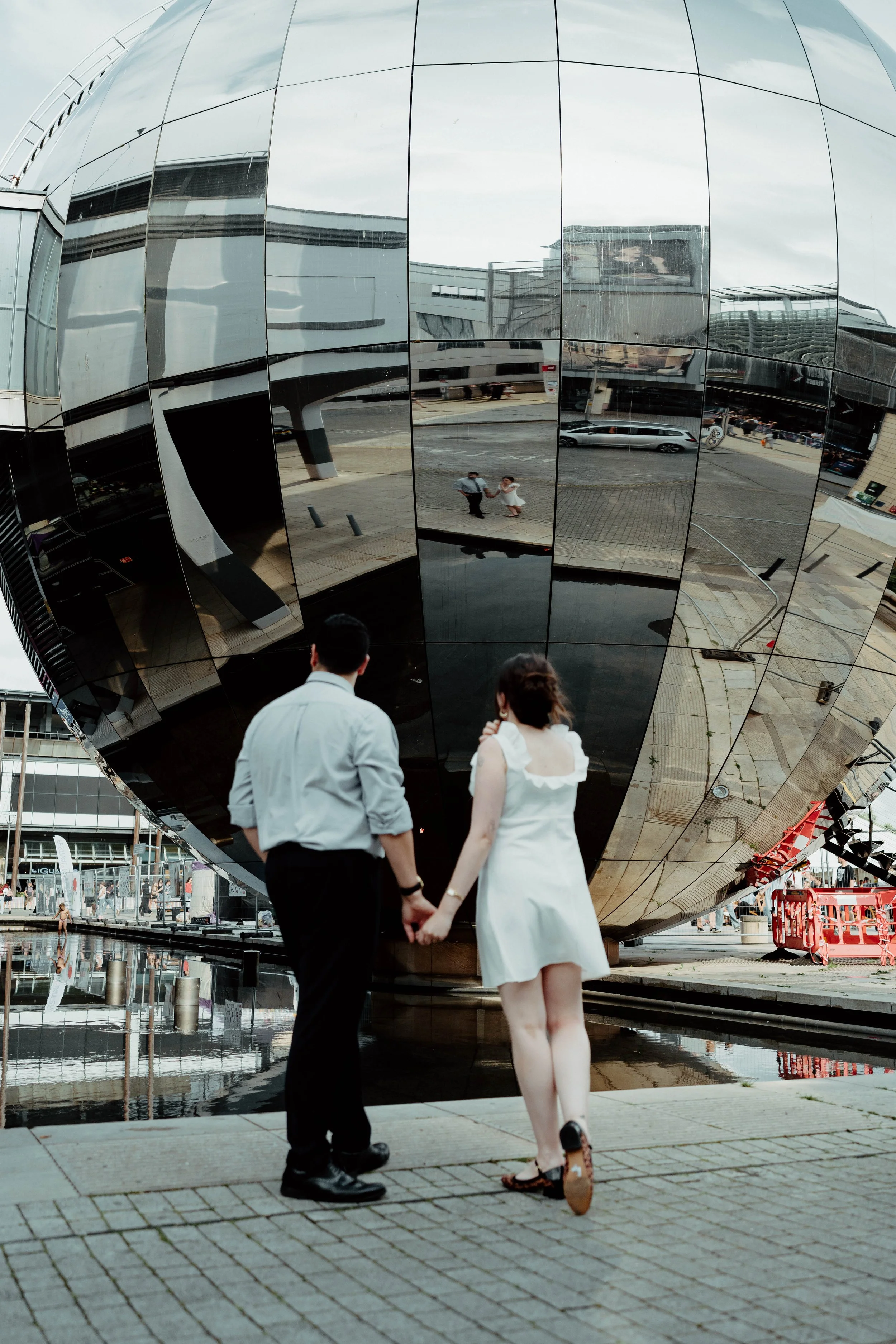 A couple holding hands outside in front of a large reflective spherical building, with their reflection visible in the sphere and the building's surface.