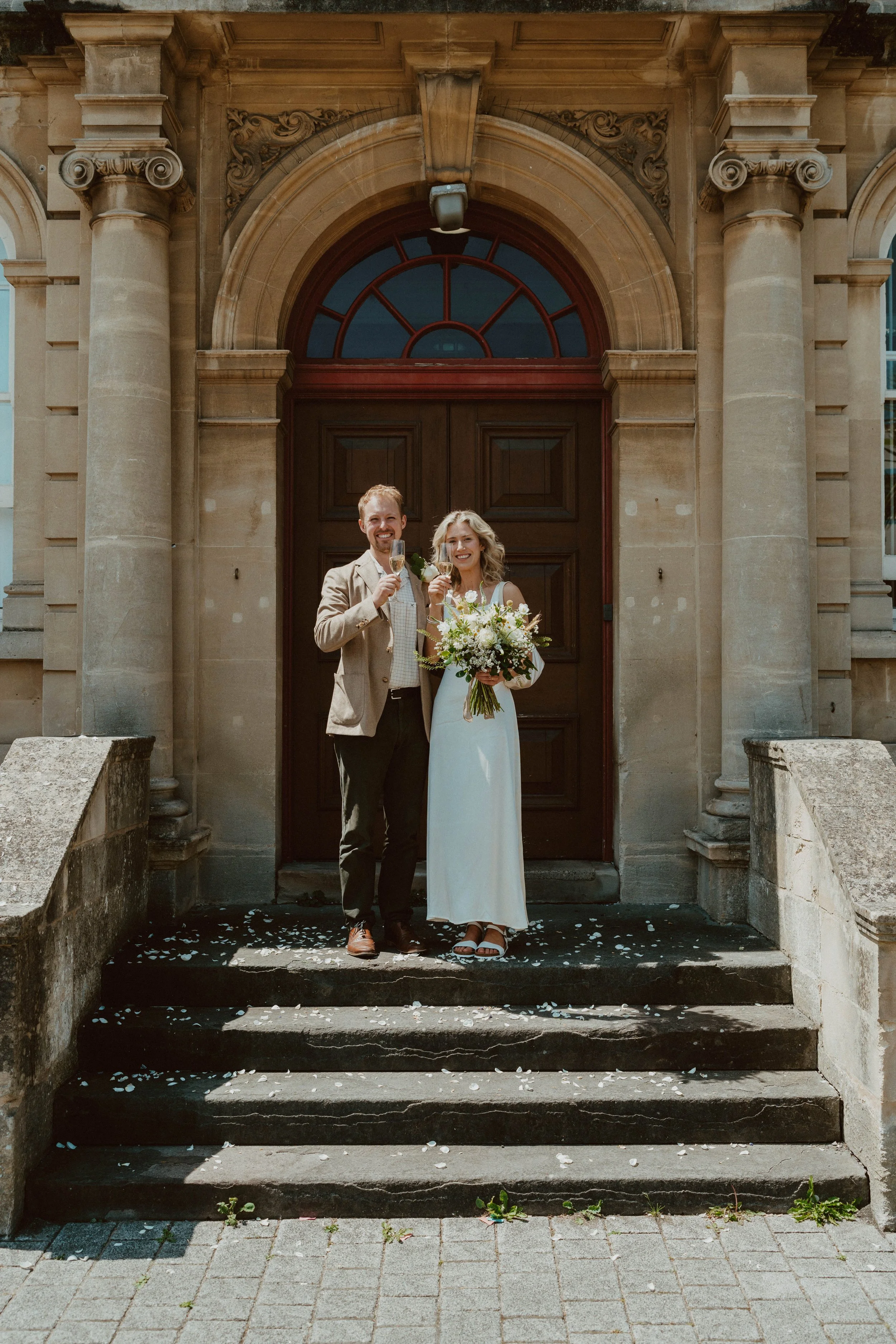A bride and groom standing on steps in front of a historic building, celebrating with glasses of champagne, with the bride holding a bouquet of flowers.