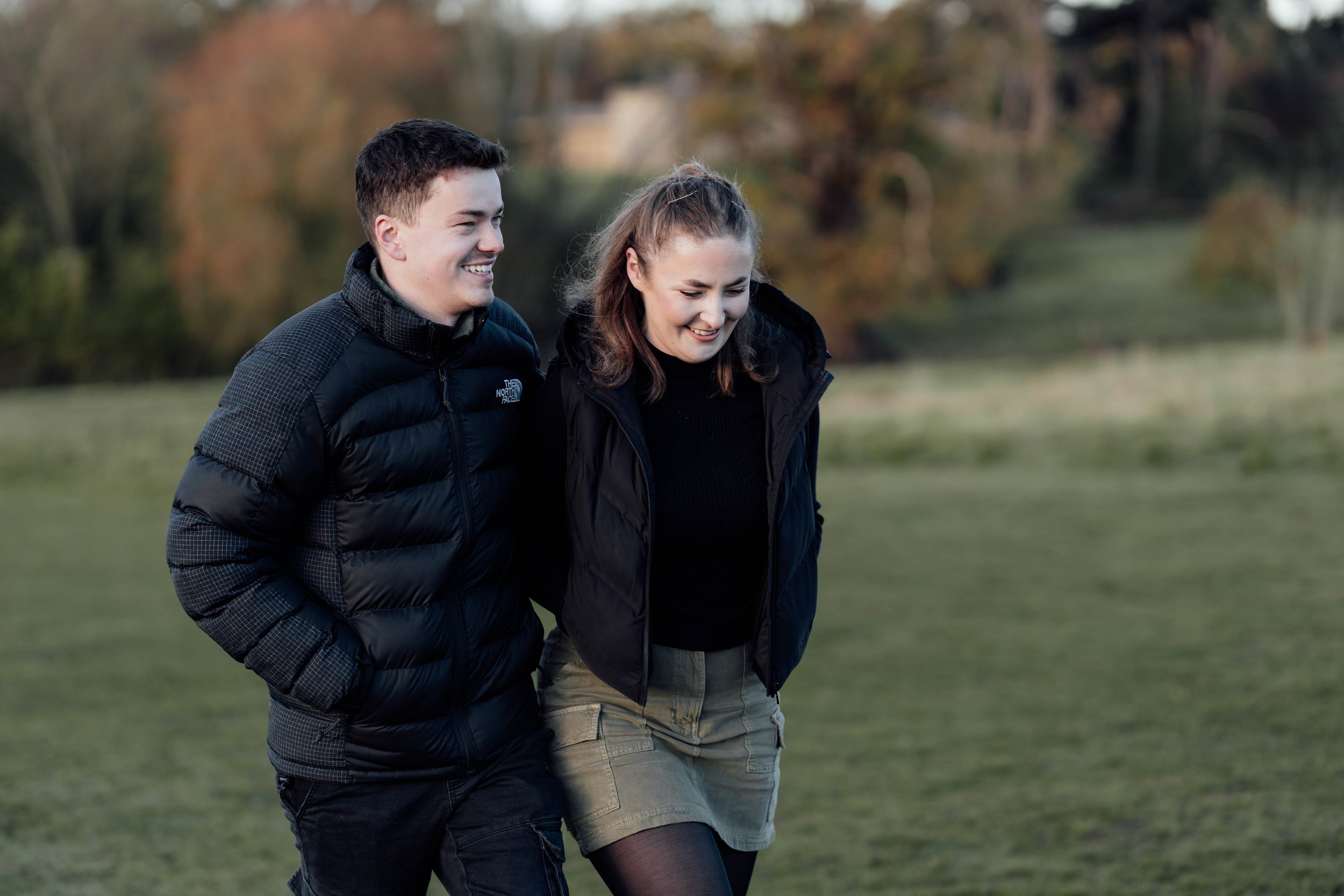 A young man and woman walking outdoors on a grassy field with trees in the background. They are smiling and wearing jackets.