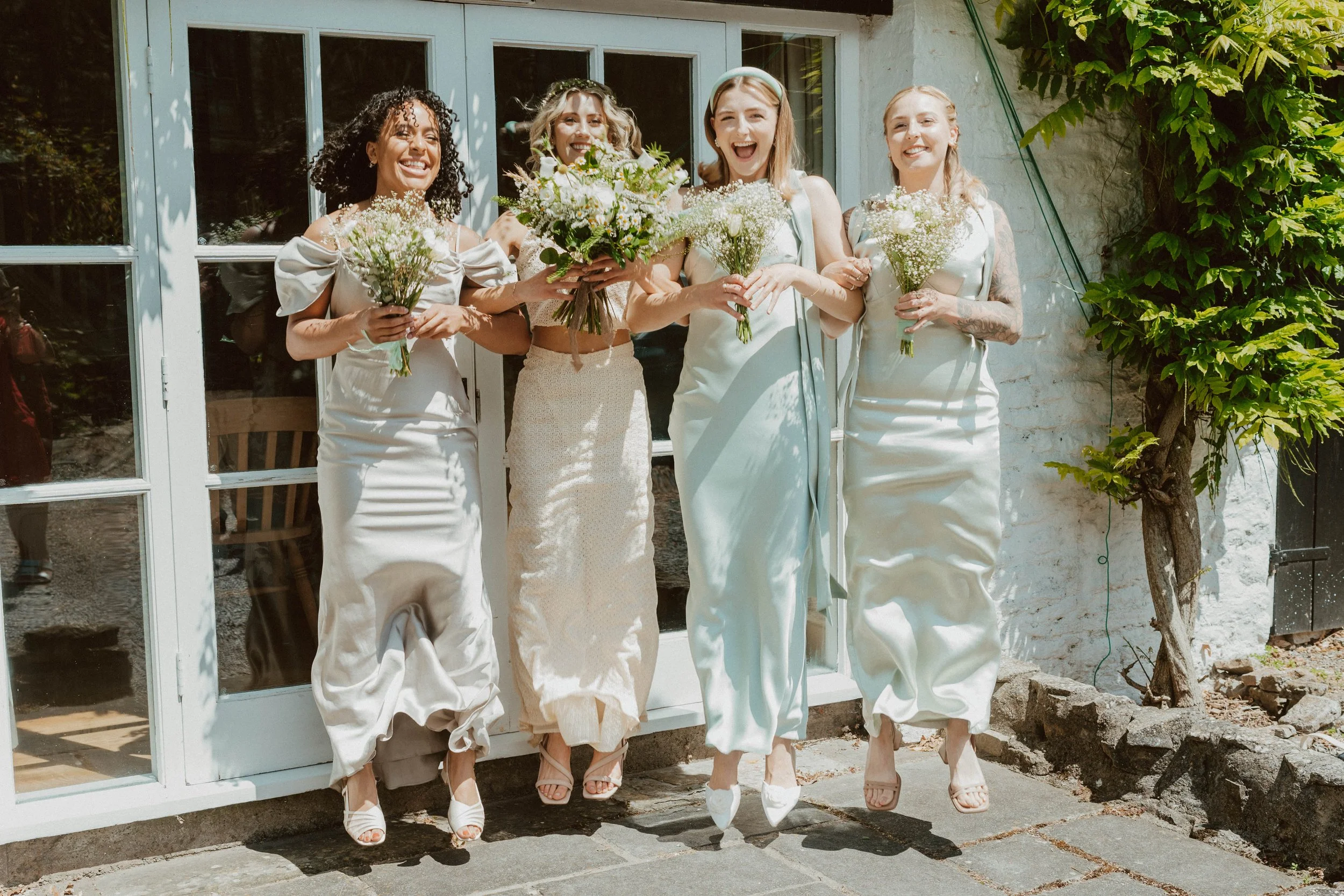 Four women in white dresses holding bouquets of white flowers, smiling, standing outside in front of a white door with glass panes.