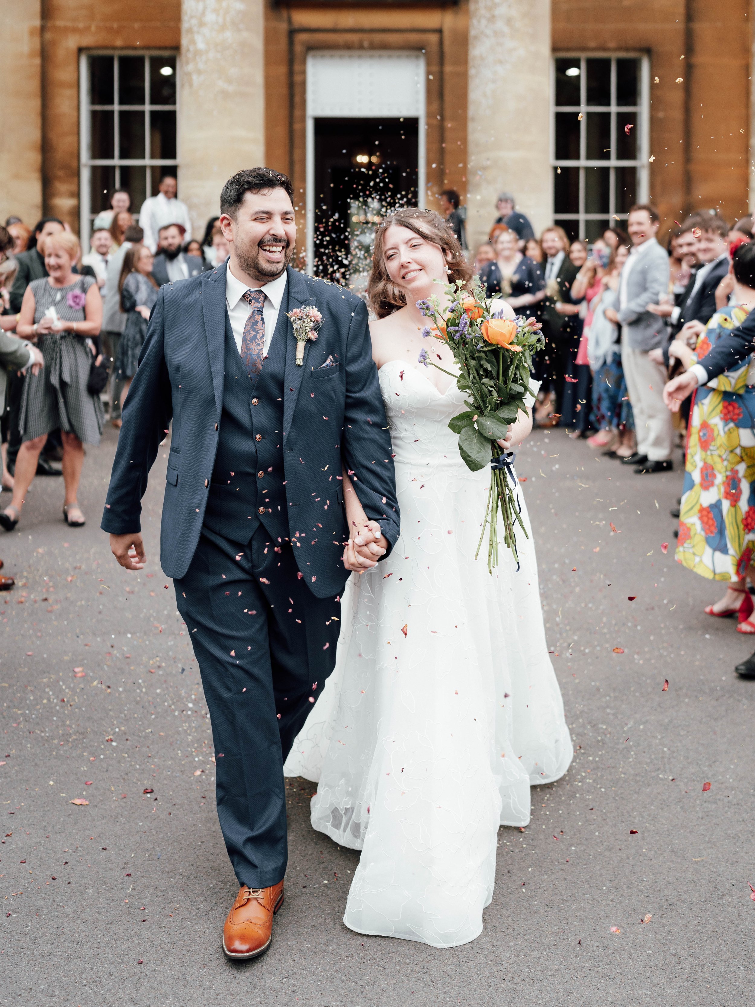 A newlywed couple walking hand in hand outside a church, smiling and surrounded by friends and family throwing confetti, during daytime.