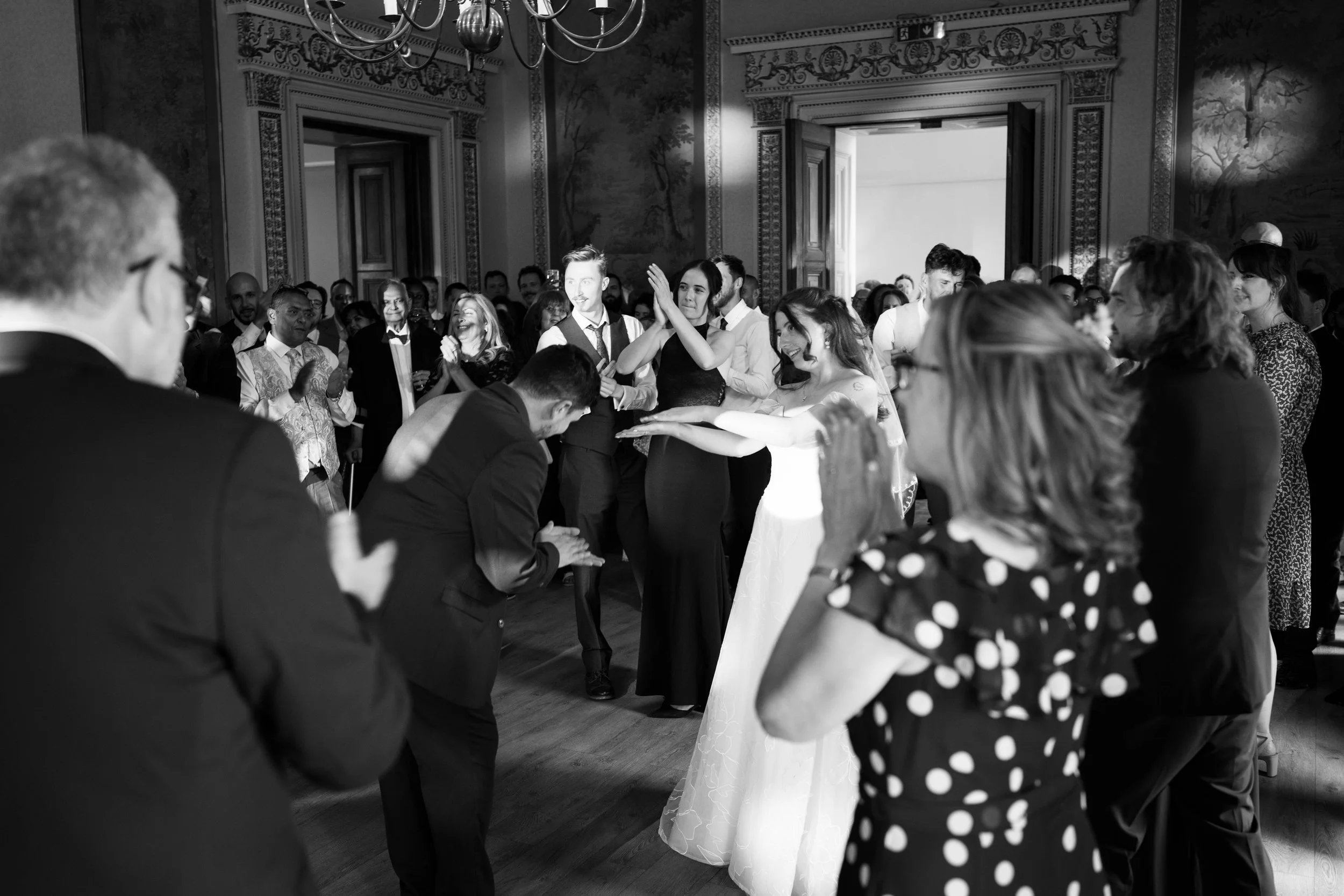 A black-and-white photo of a wedding reception with guests dancing and enjoying themselves in an ornate hall.