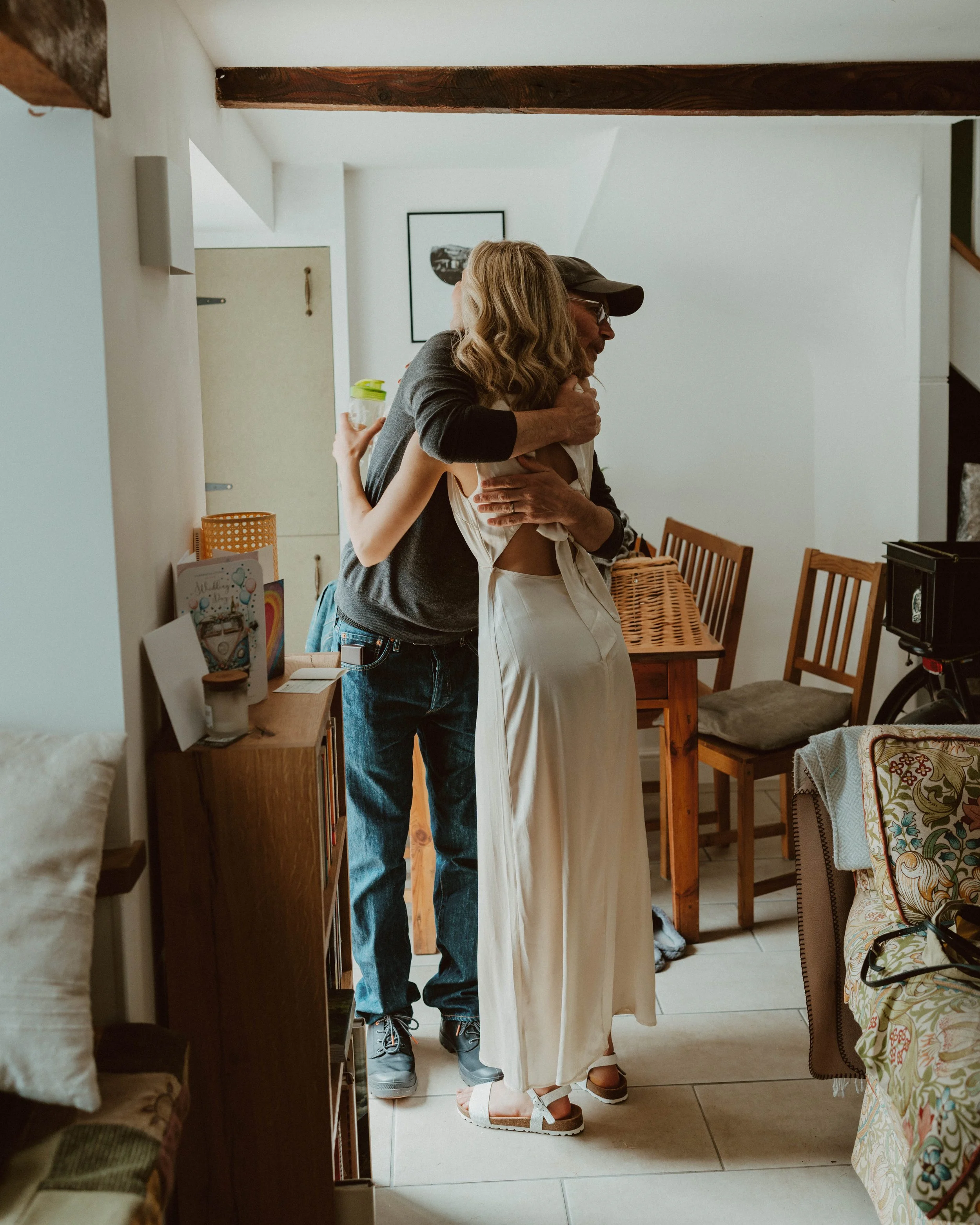 Two people hugging in a cozy indoor setting, one wearing a white dress and the other in a dark shirt and jeans.