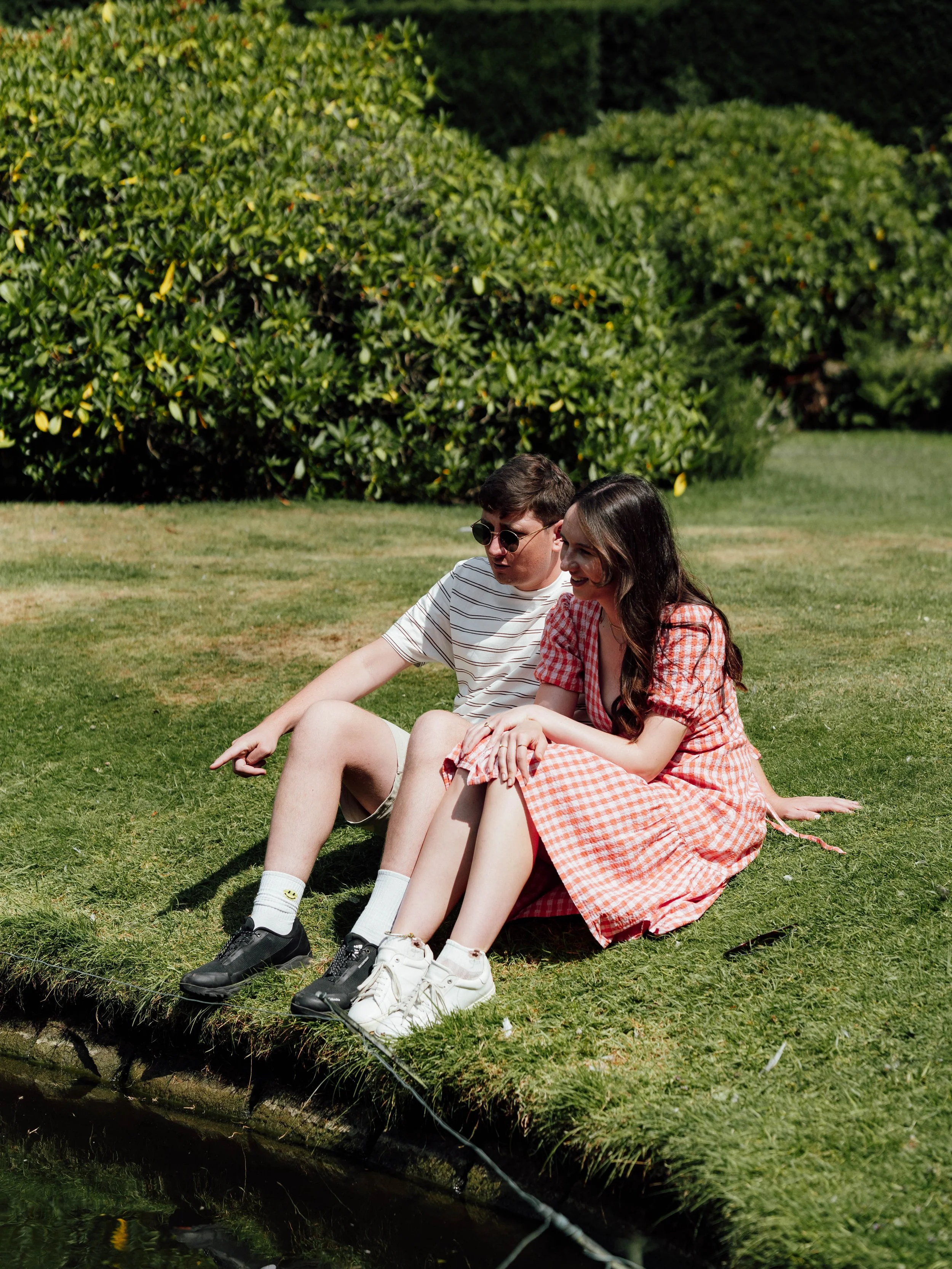 A young man and woman sitting on grass near a small pond, smiling and enjoying a sunny day outside.