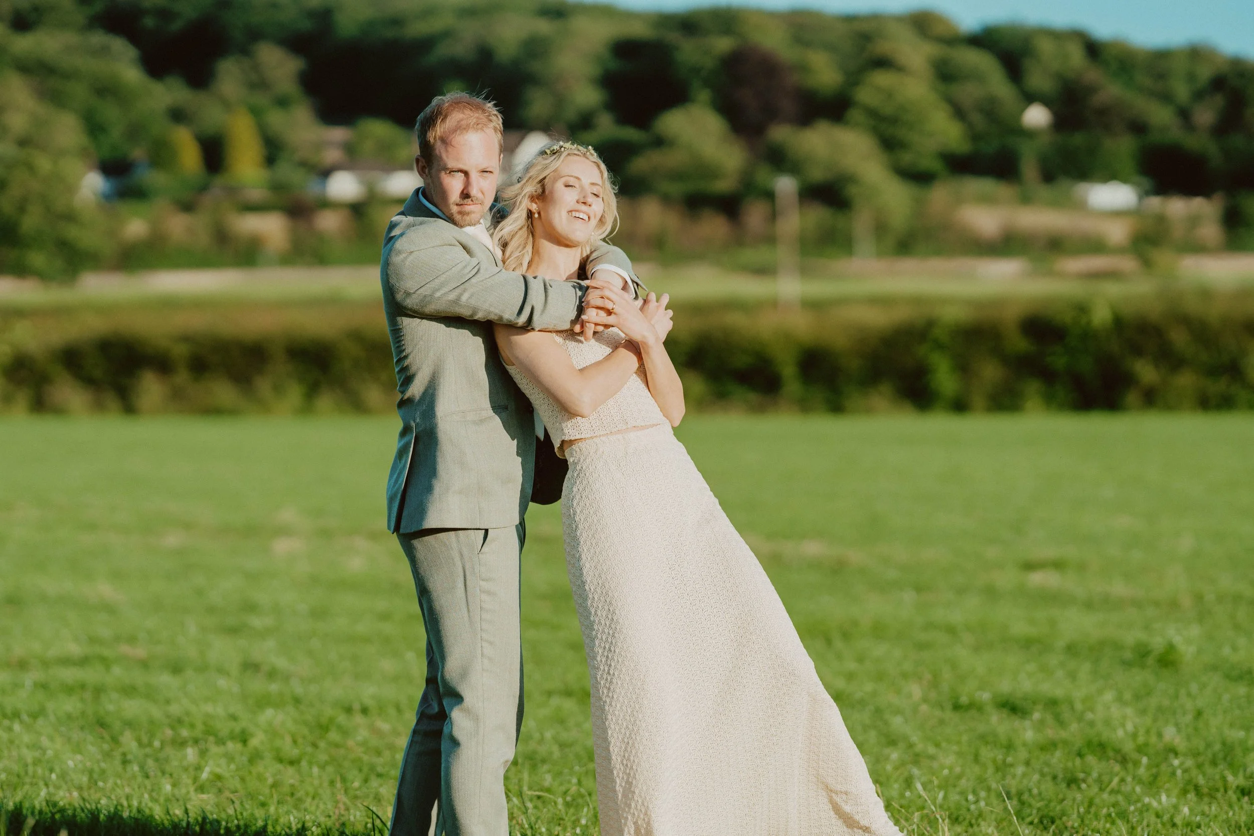 A man and woman in wedding attire embracing outdoors on a grassy field with trees and hills in the background.