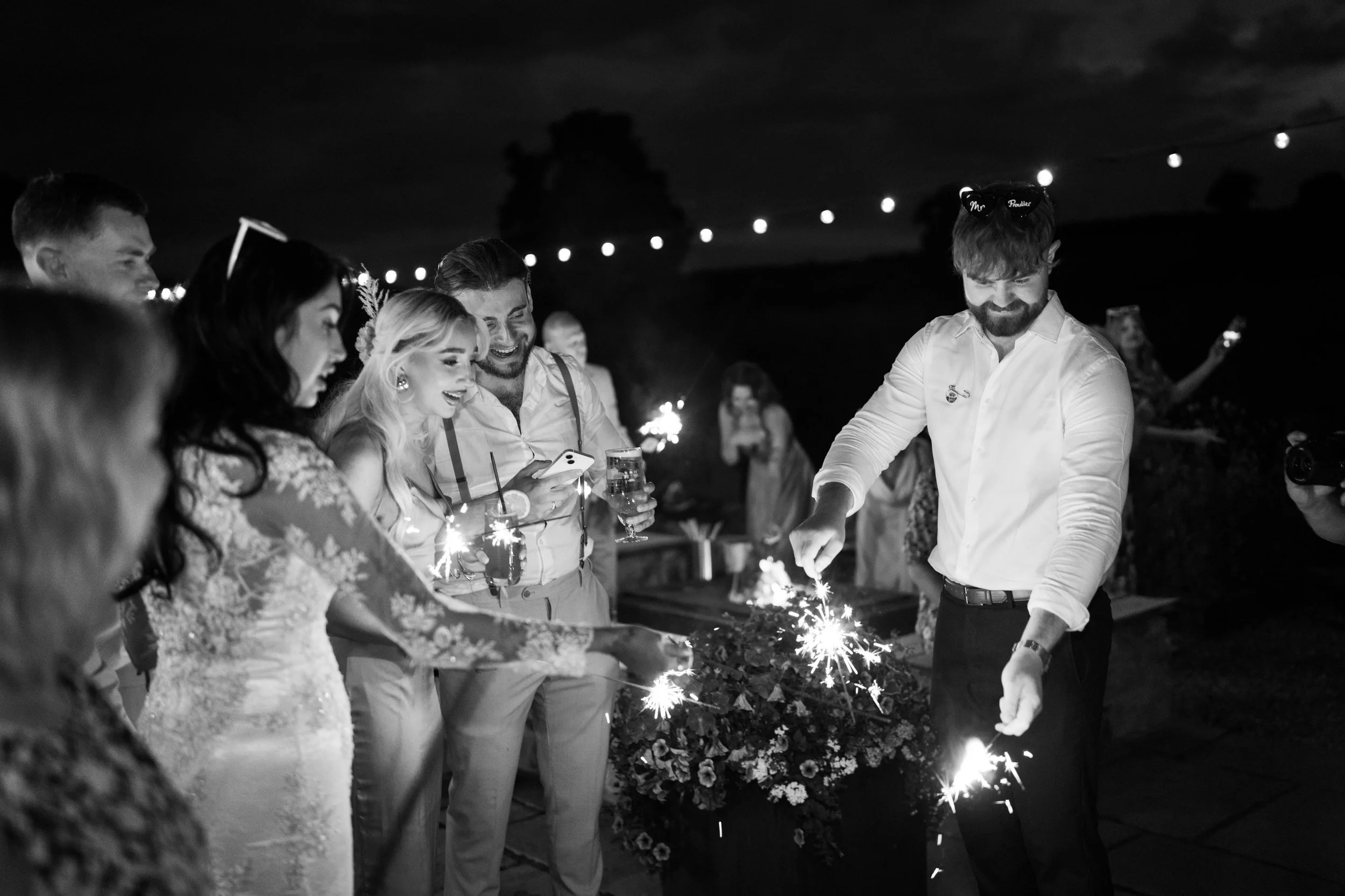 Group of people celebrating at a wedding reception at night, holding sparklers and drinks, smiling, with string lights in the background.