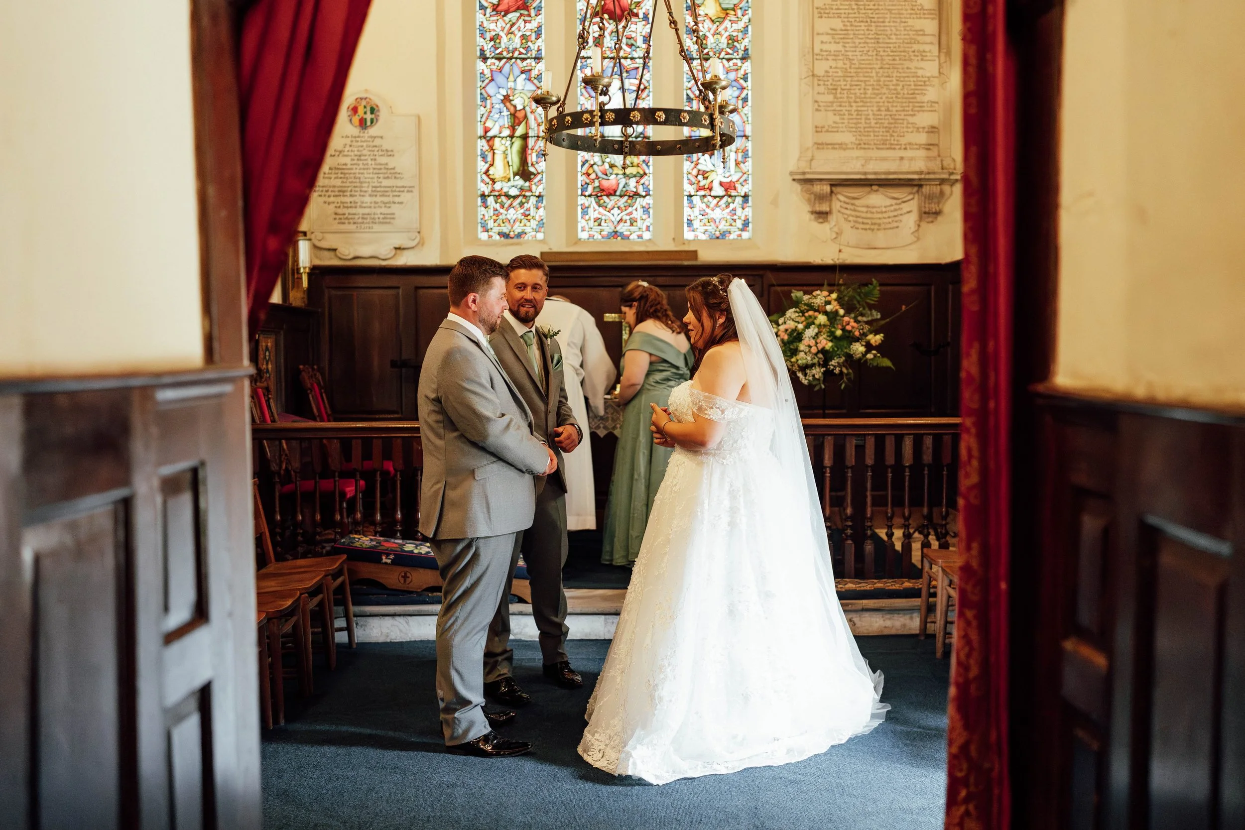 A bride and groom exchanging vows in a church with stained glass windows, while a woman in a green dress stands behind them. The bride wears a white wedding gown and veil, and the groom wears a gray suit. Red curtains frame the view.