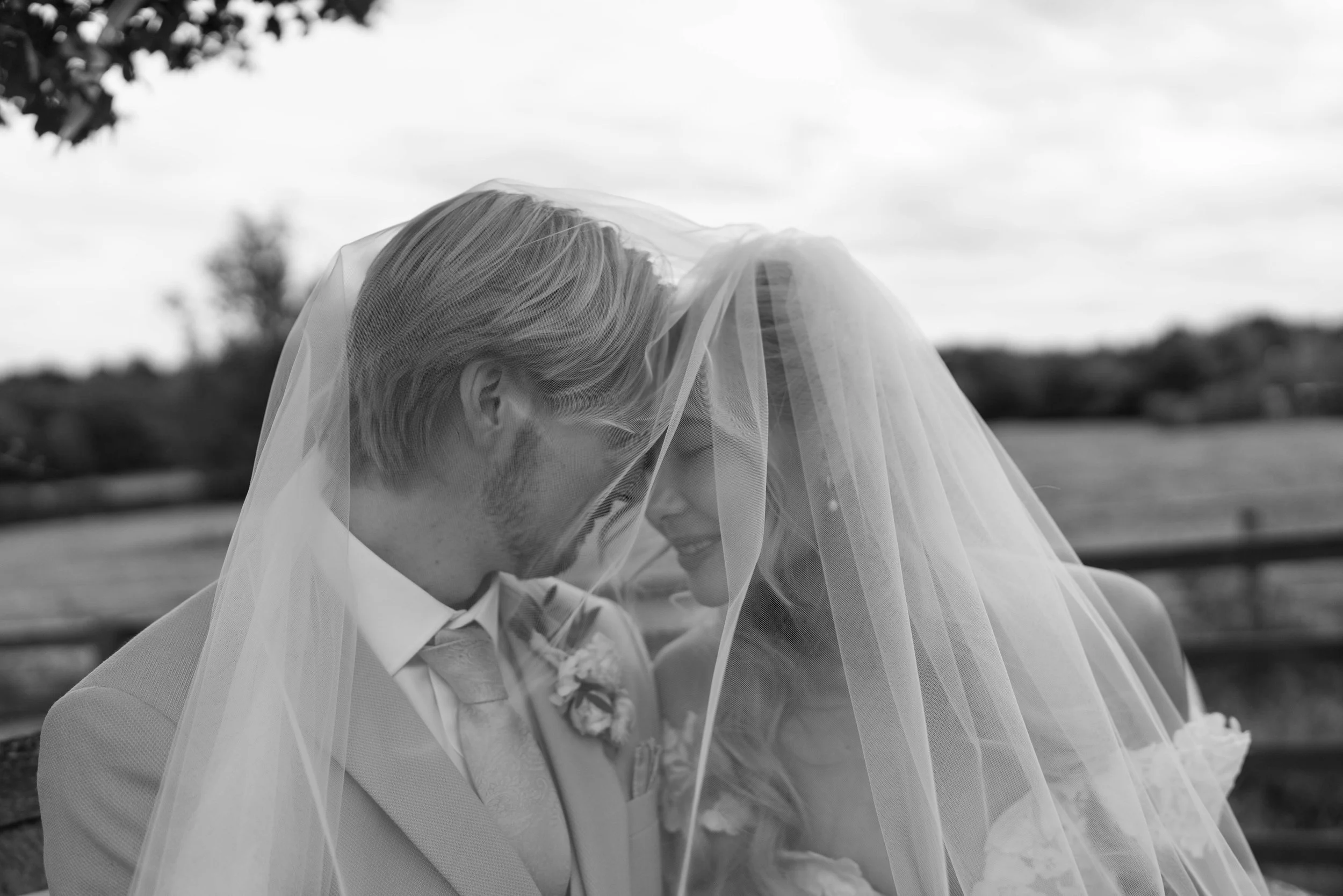 Black and white photo of a bride and groom smiling under a wedding veil, outdoors with a wooden fence and cloudy sky in the background.