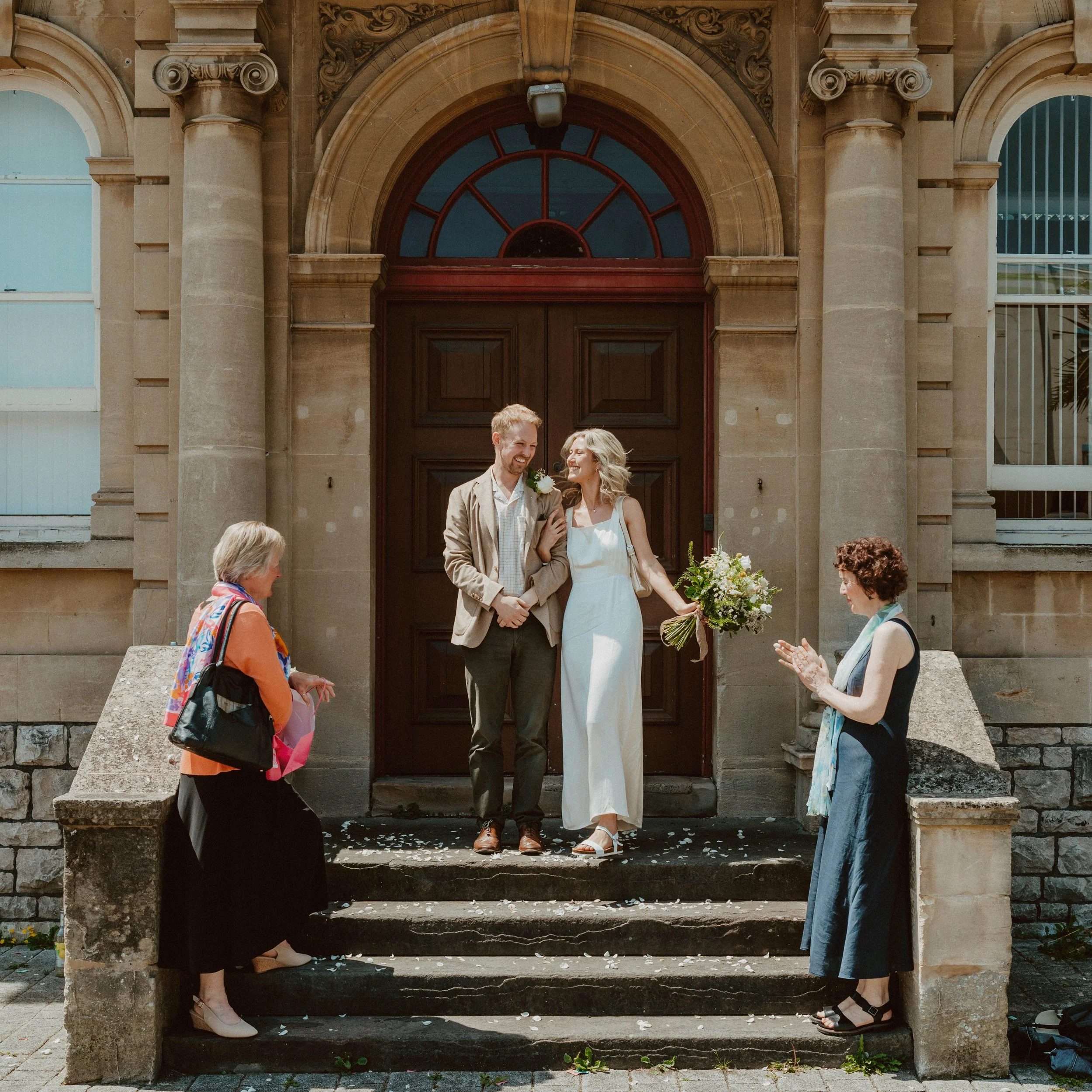 A happy couple standing on the steps of a historic building for a wedding celebration. The man is wearing a beige blazer and the woman is in a white dress holding a bouquet. Two women are clapping and celebrating beside them.