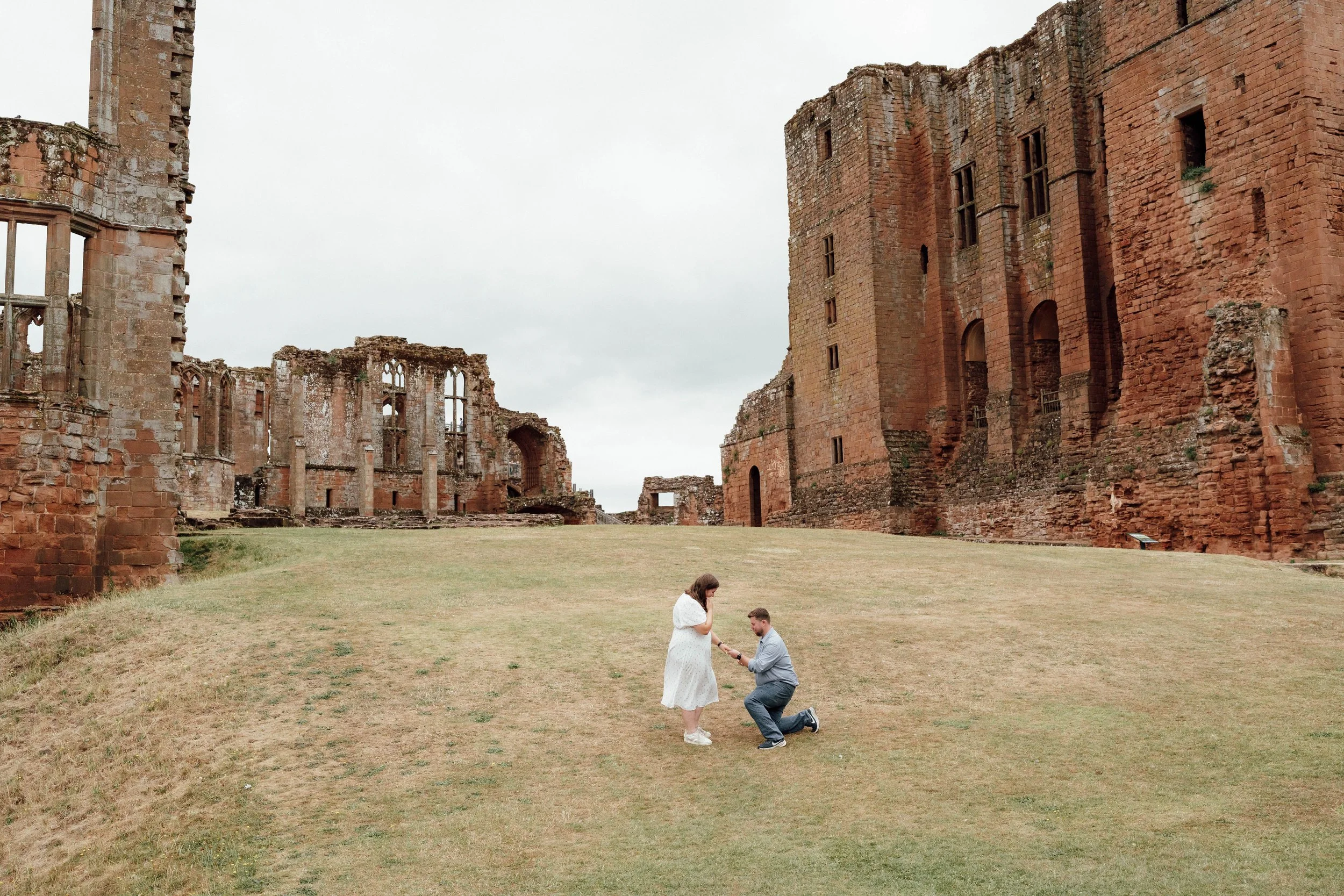 A man is proposing marriage to a woman in front of the ruins of an ancient castle. The woman is wearing a white dress and is holding the man's hand, which has a ring. They are standing on a grassy area with an overcast sky.