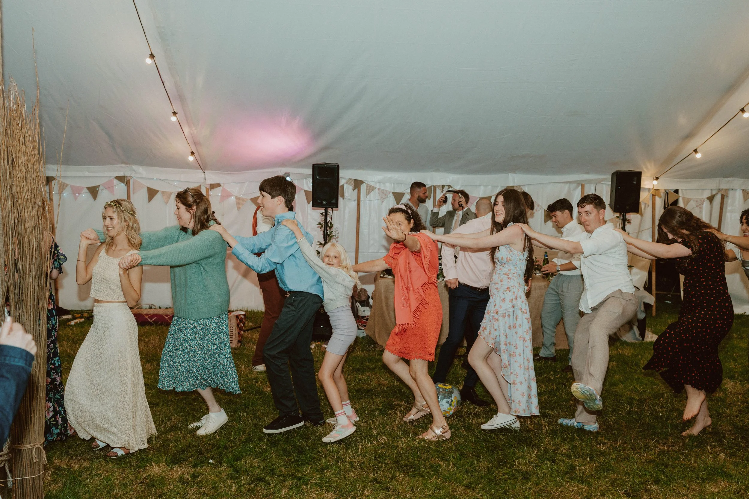 A group of people, including children and adults, participating in a conga line dance inside a decorated tent at a celebration or wedding reception, with string lights and bunting decorations overhead.