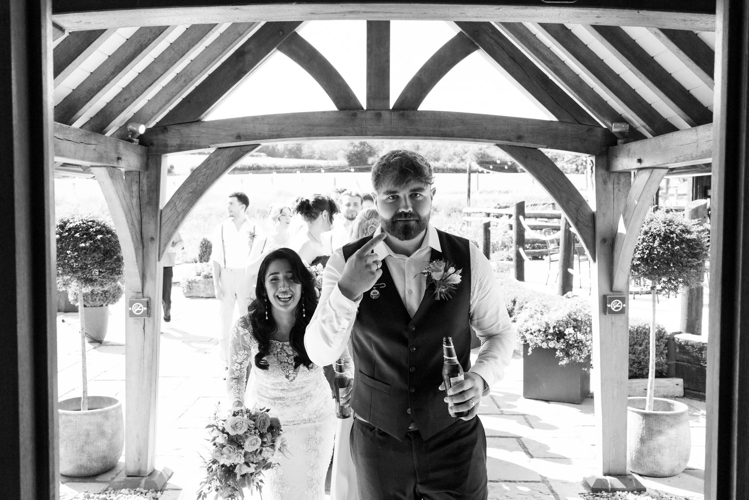 Black and white photo of a wedding reception, with a man in a vest and white shirt holding a drink and making a gesture with his hand, and smiling woman in a wedding dress holding a bouquet. Other guests are in the background.