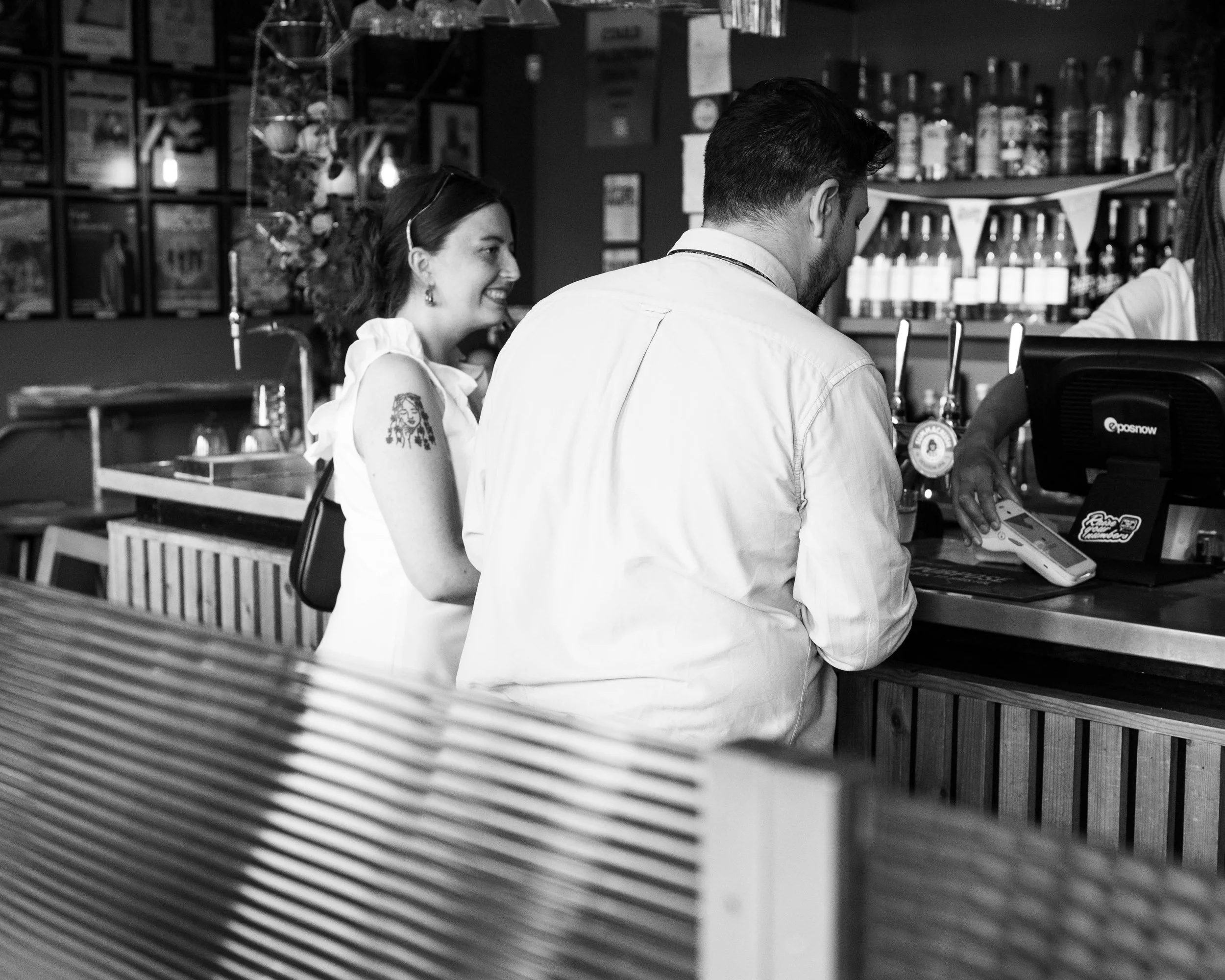 A woman with a tattoo on her arm smiling at a man at a bar counter in a cafe or bar, with shelves of bottles and framed pictures on the wall in the background.