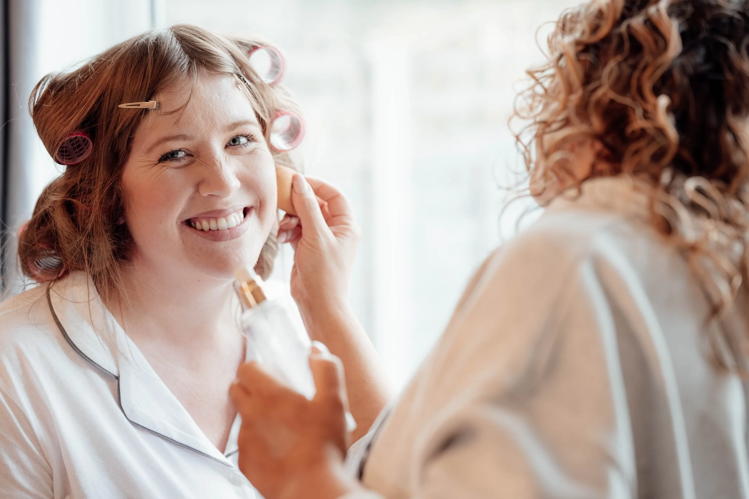 A woman with curlers in her hair smiling as she gets her makeup done.
